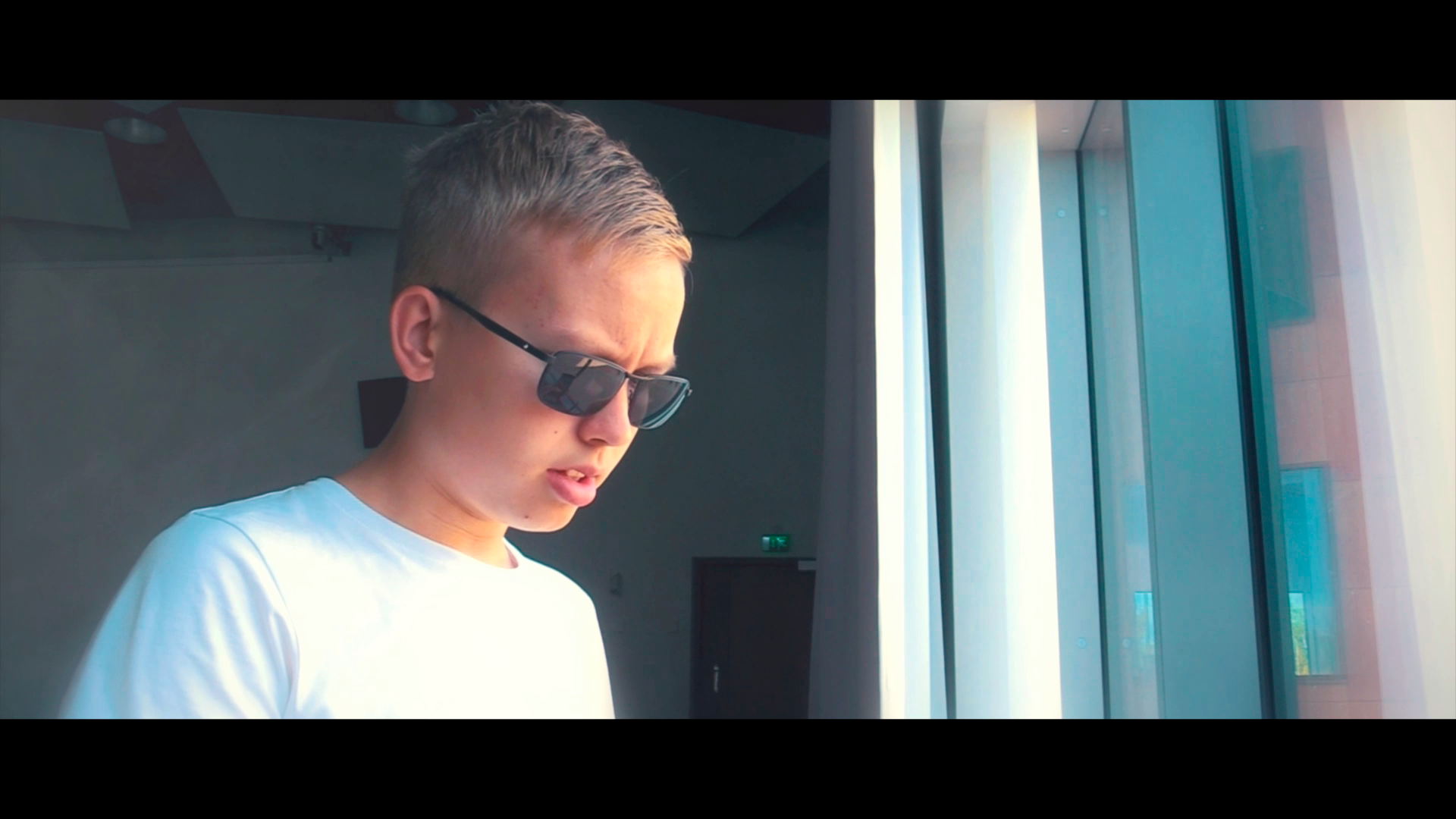 Nuori mies katsoo ulos ikkunasta, pukeutunut valkoiseen t-paitaan ja käyttää tummia aurinkolaseja. Charles Smith - Tehtävä Kangasalla -elokuva, kuvakaappaus elokuvasta, Media Aaro, Aaro Riponiemi