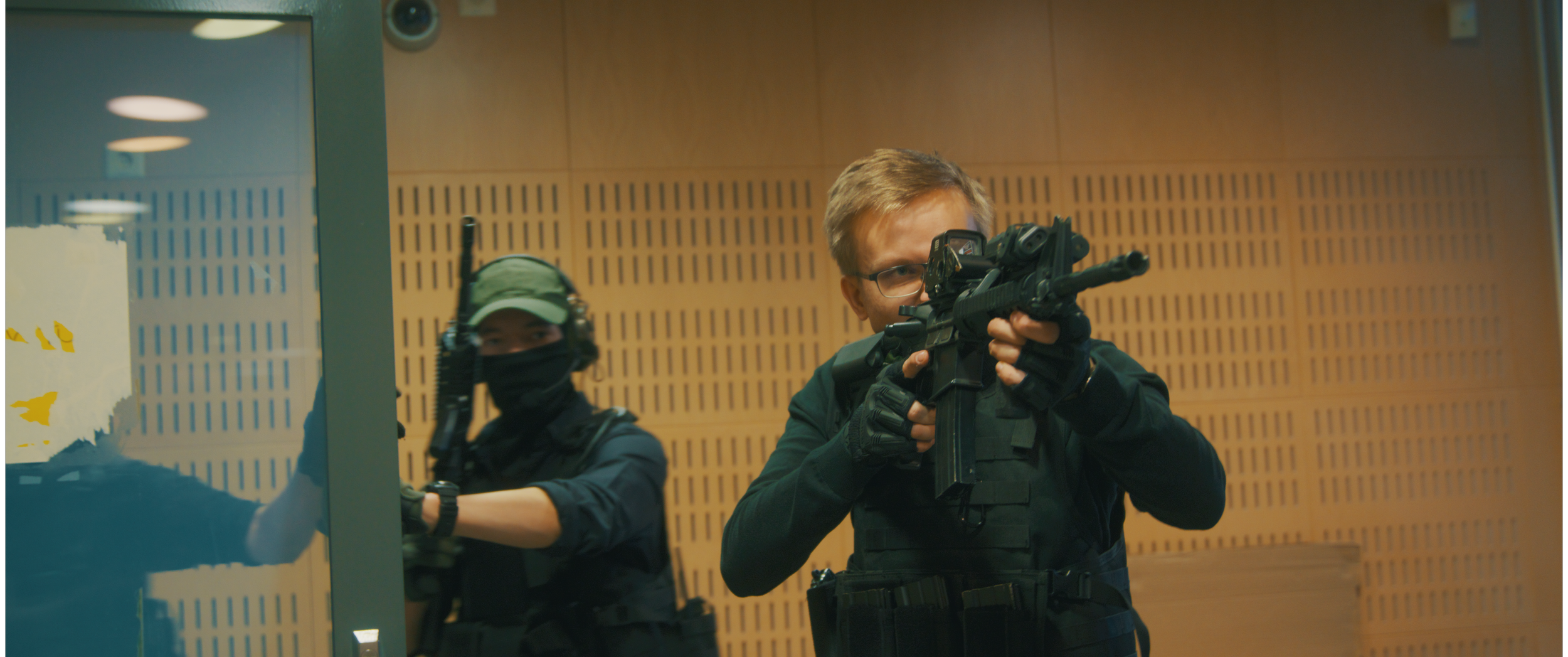 Kaksi aseistettua poliisia harjoittelee ampujaa sisätiloissa, toinen ammuskääntään tähtäintä ja toinen on suojaamassa sivulla, Charles Smith - Koston Kyynel -elokuva, kuvakaappaus elokuvasta, Media Aaro, Aaro Riponiemi