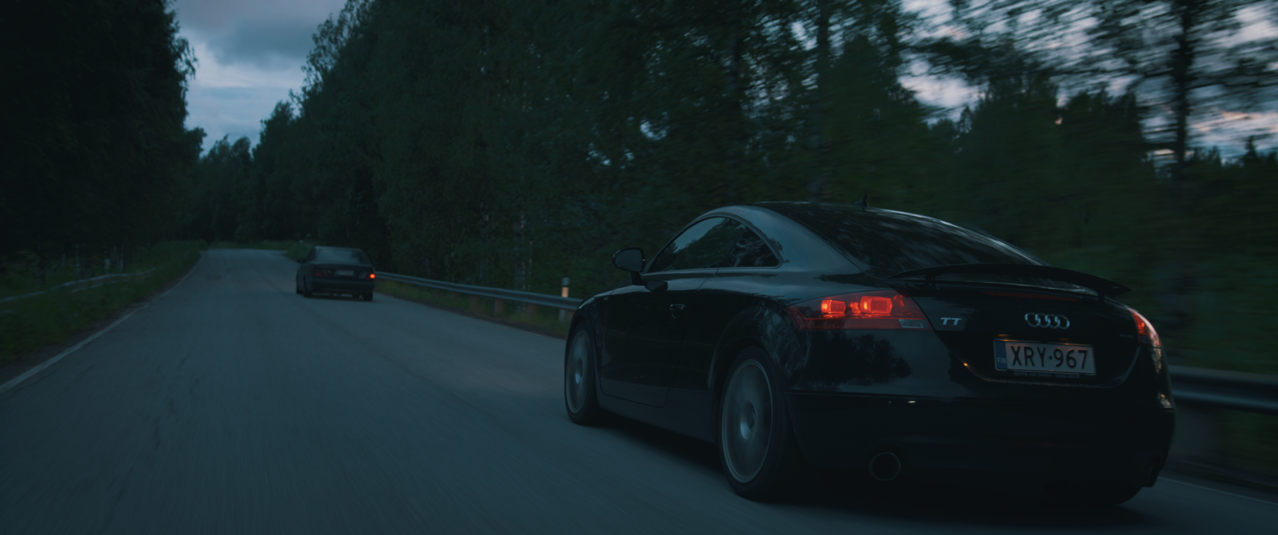 Musta Audi TT ajamassa kapealla maantiellä metsän keskellä iltahämärässä, Charles Smith - Koston Kyynel -elokuva, kuvakaappaus elokuvasta
