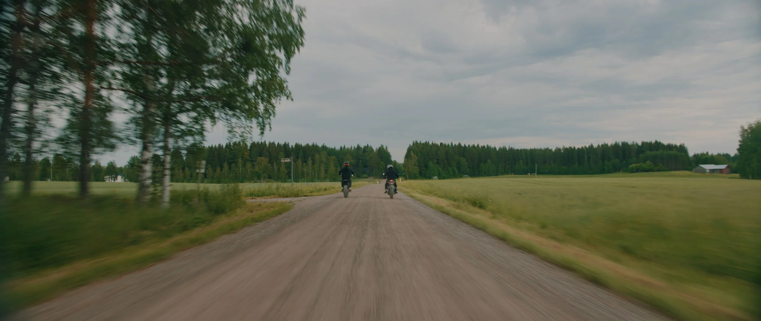 Kaksi motoristia ajavat kylätietä maaseudulla, ympärillä vihreitä puita ja niittyjä, pilvetön taivas, Charles Smith - Koston Kyynel -elokuva, kuvakaappaus elokuvasta