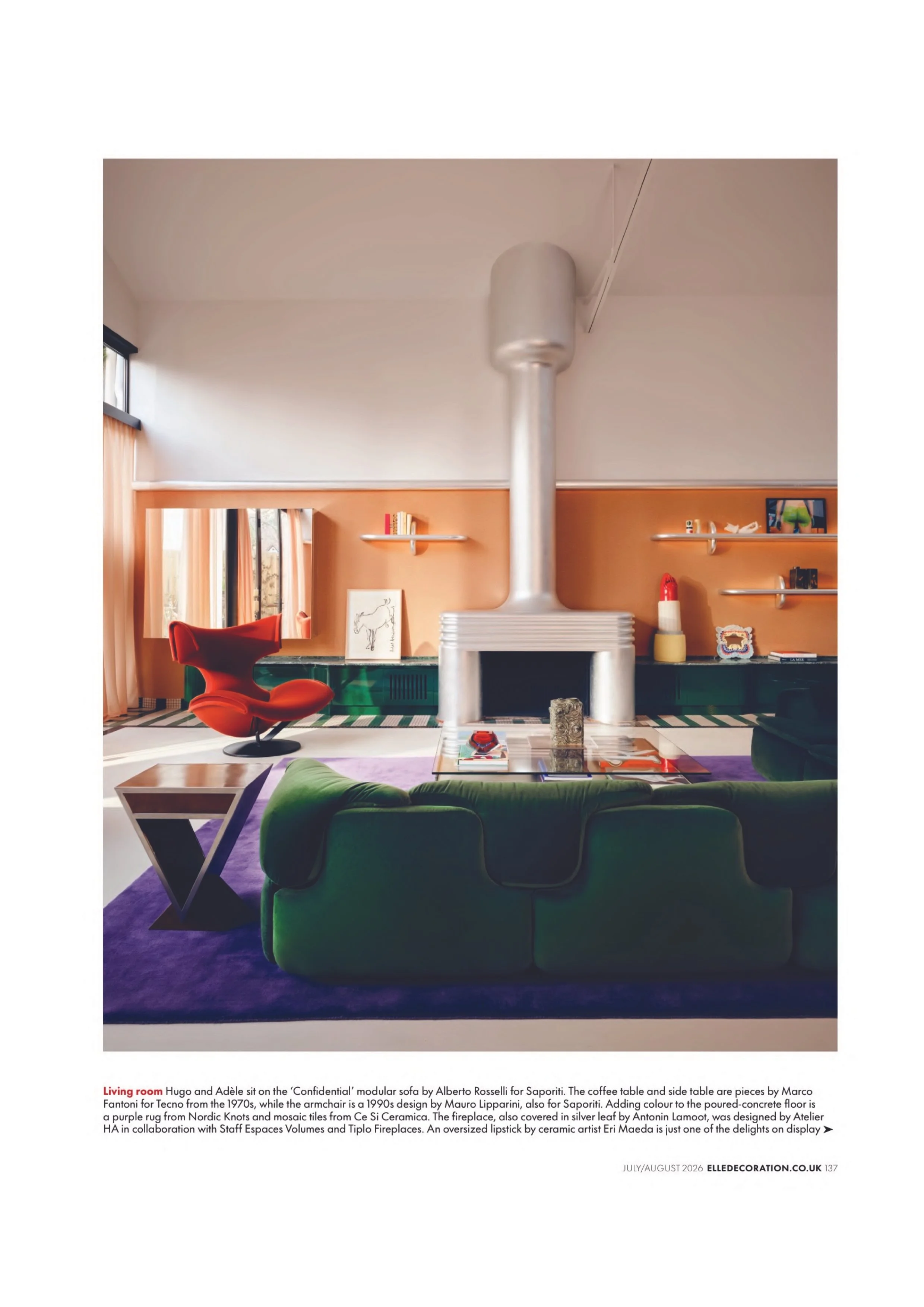 Living room with a silver central fireplace, orange and black furniture, and decorative shelves with artwork and objects, accented by patterned carpet and large windows.