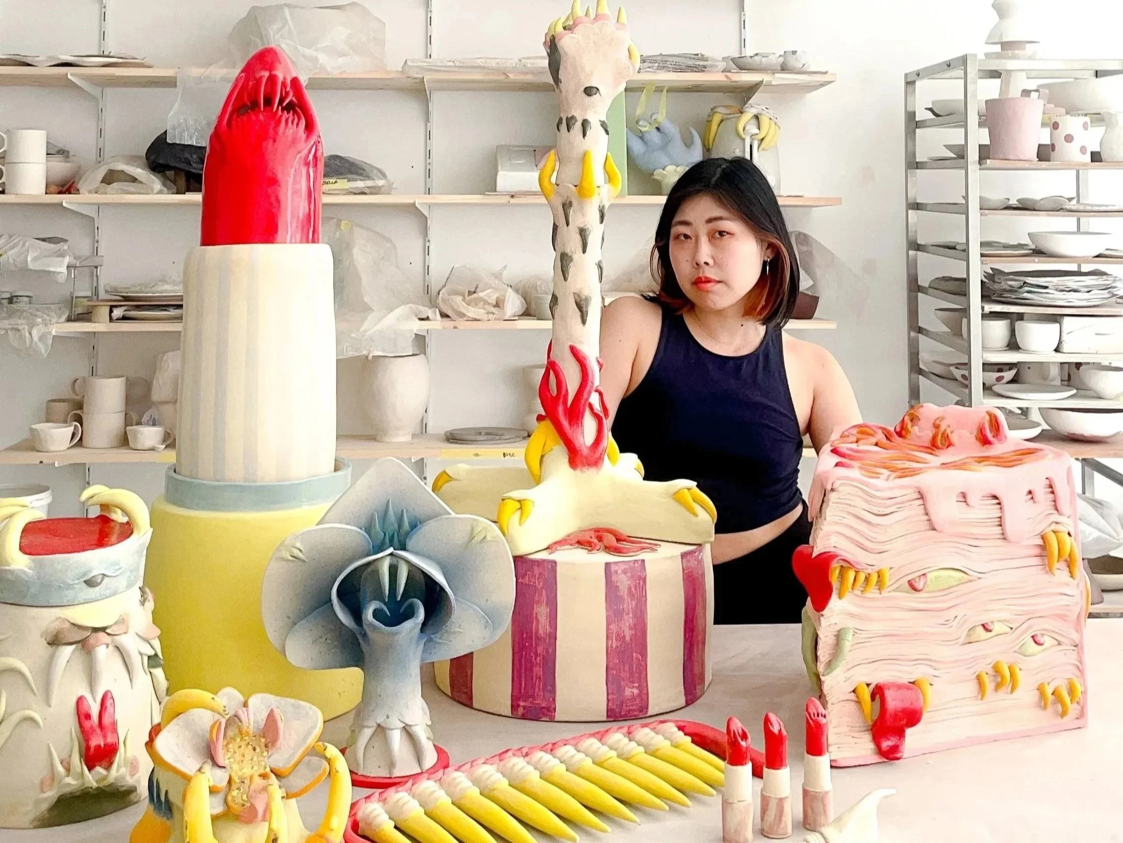 A woman with dark hair and wearing a black sleeveless top is seated in front of colorful ceramic sculptures and pottery in an art studio with shelves of finished pottery in the background.