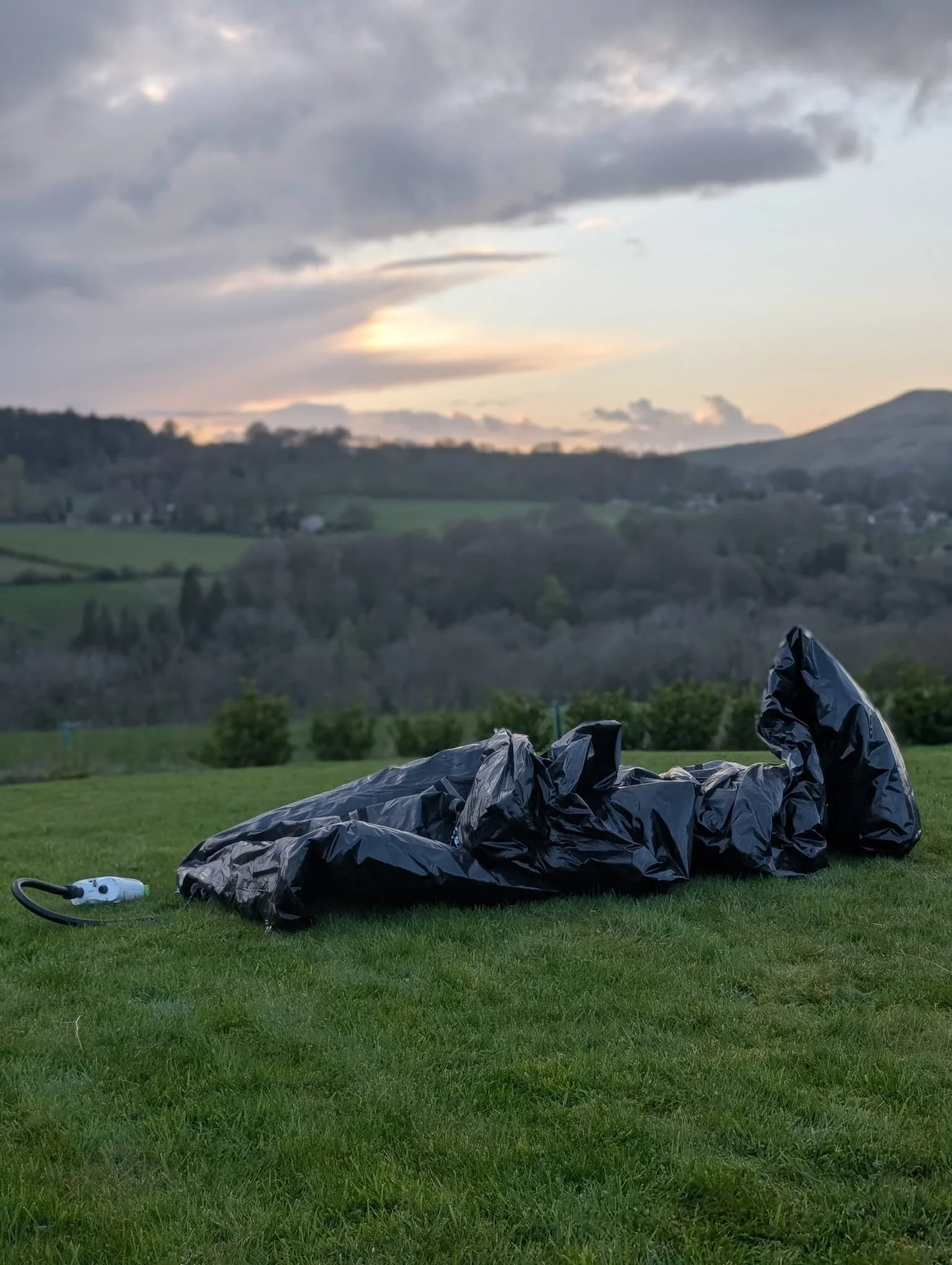 A black trash bag on green grass with a cloudy sky and distant hills in the background