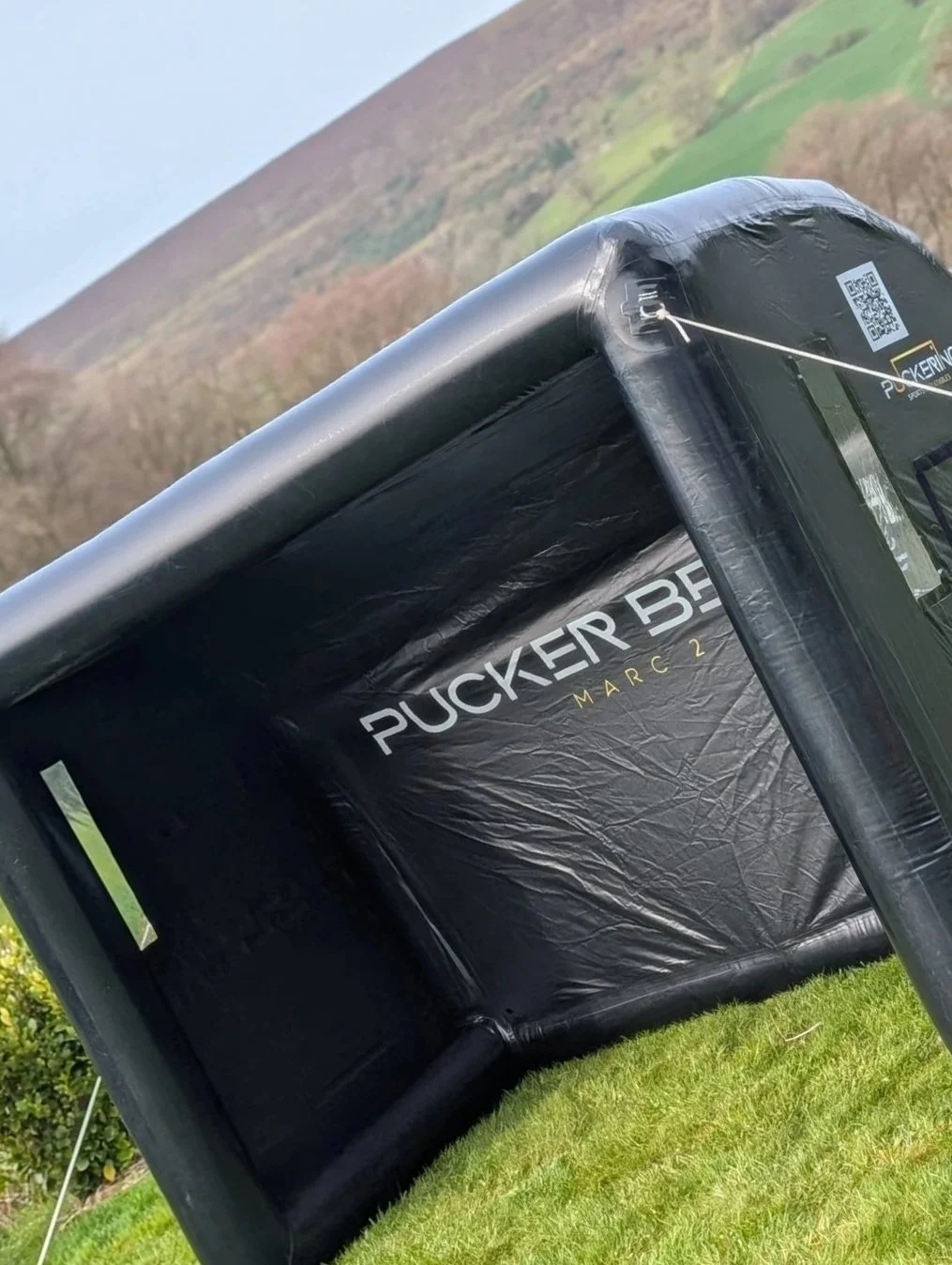 An inflatable shelter or tent labeled "Puckier Base March 2" set up on grass with hills in the background.