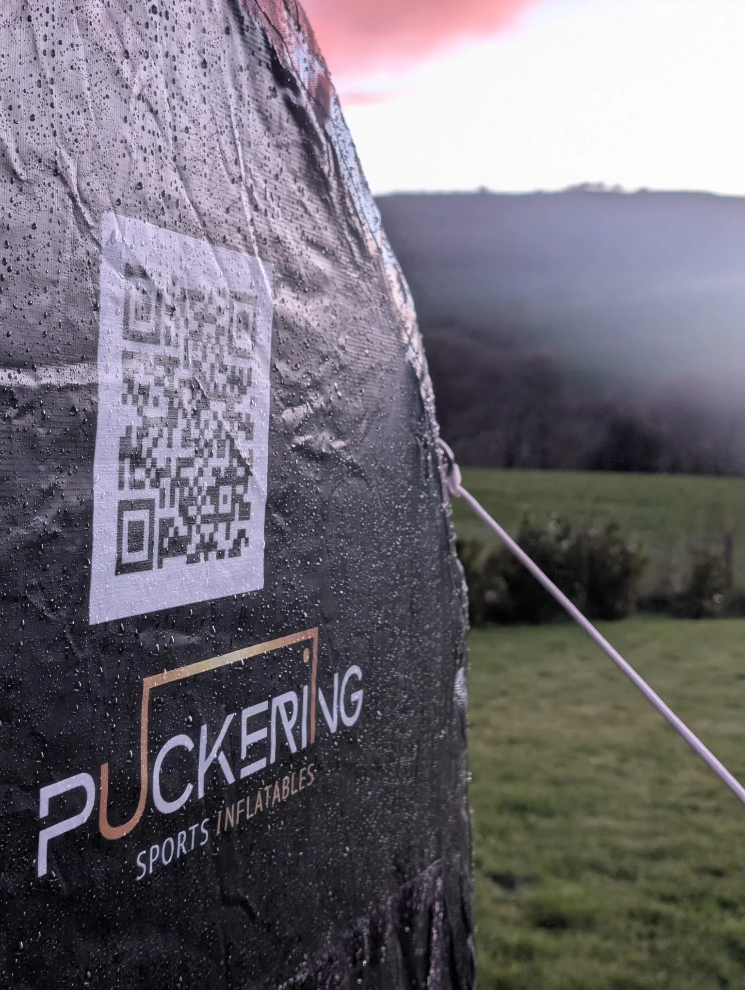 Close-up of a Deflategate sports inflatables object with water droplets, an attached QR code, and a BUCKE RING SPORTS INFLATABLES logo, set in a grassy outdoor area with trees and a mountain in the background at sunset.