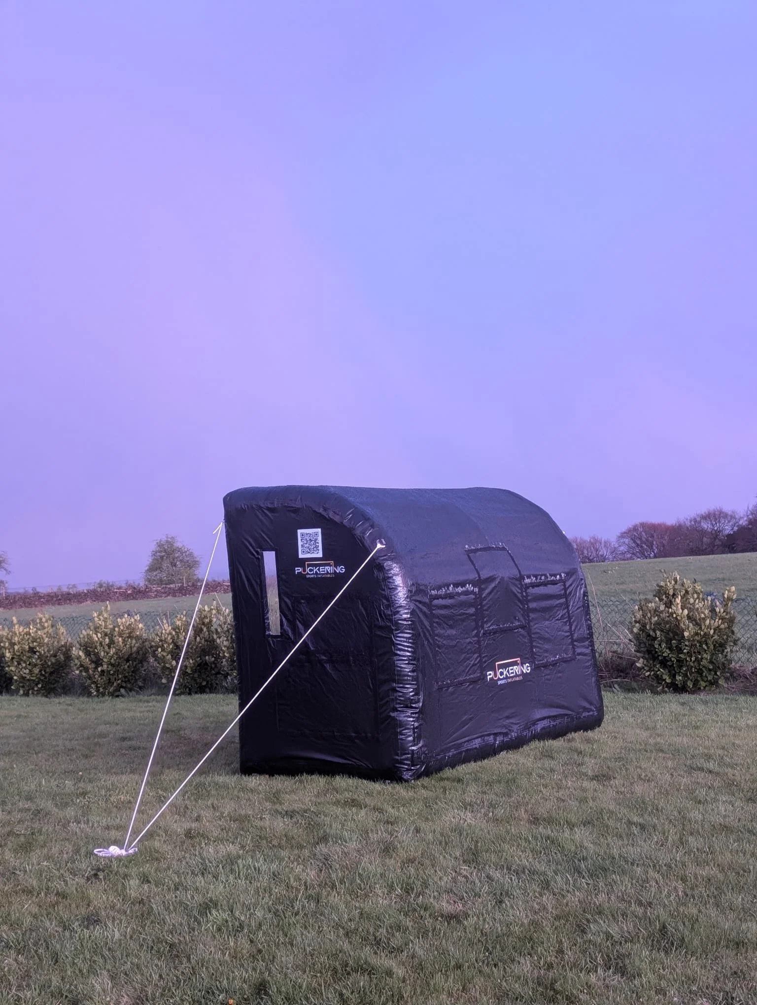 A black sports tent set up on a grassy area, with a chain-link fence and bushes in the background, during twilight or early evening.
