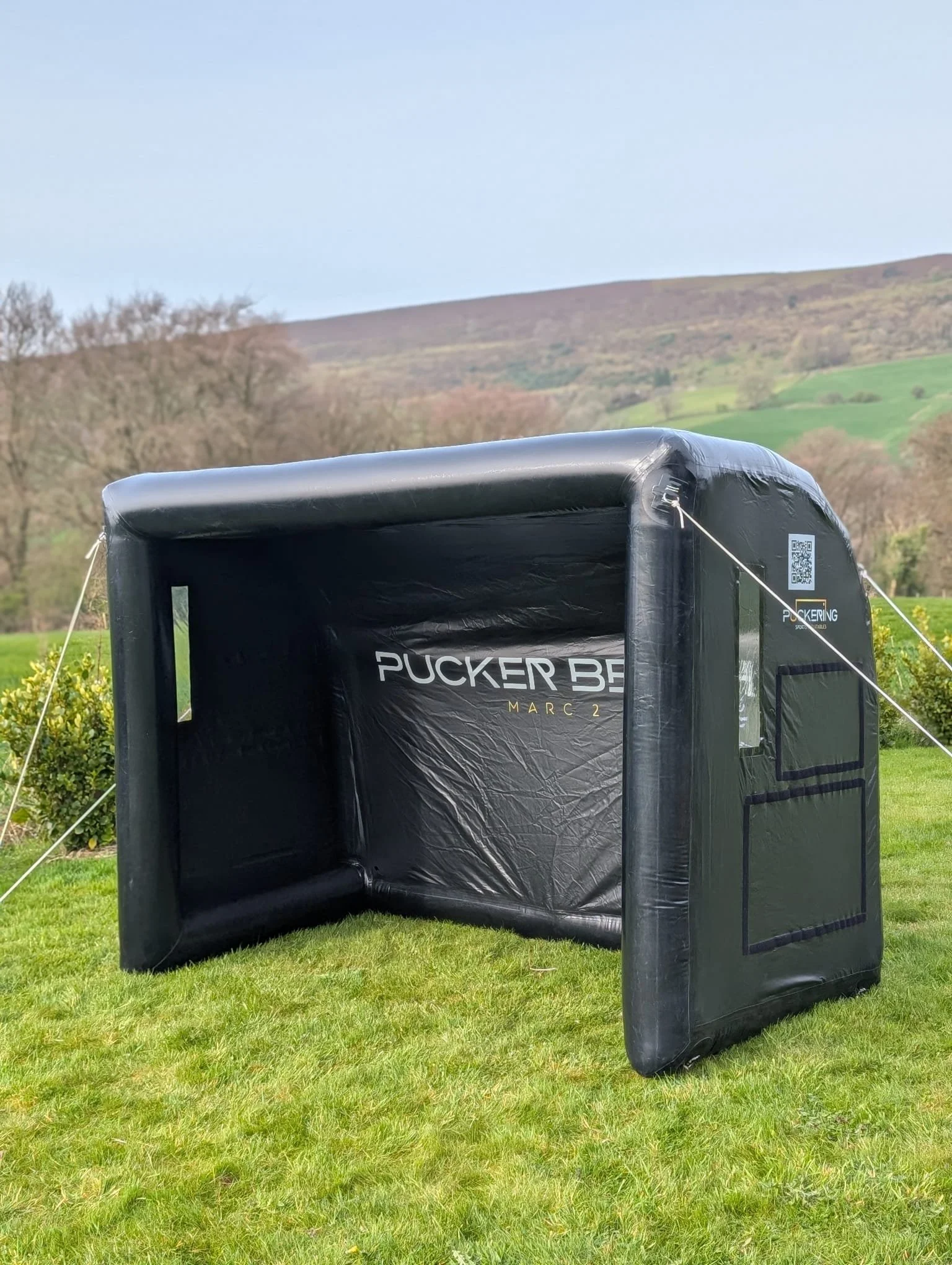 Inflatable dugout set up outdoors on grassy field with hills and trees in background, branded 'Pucker Bench'.