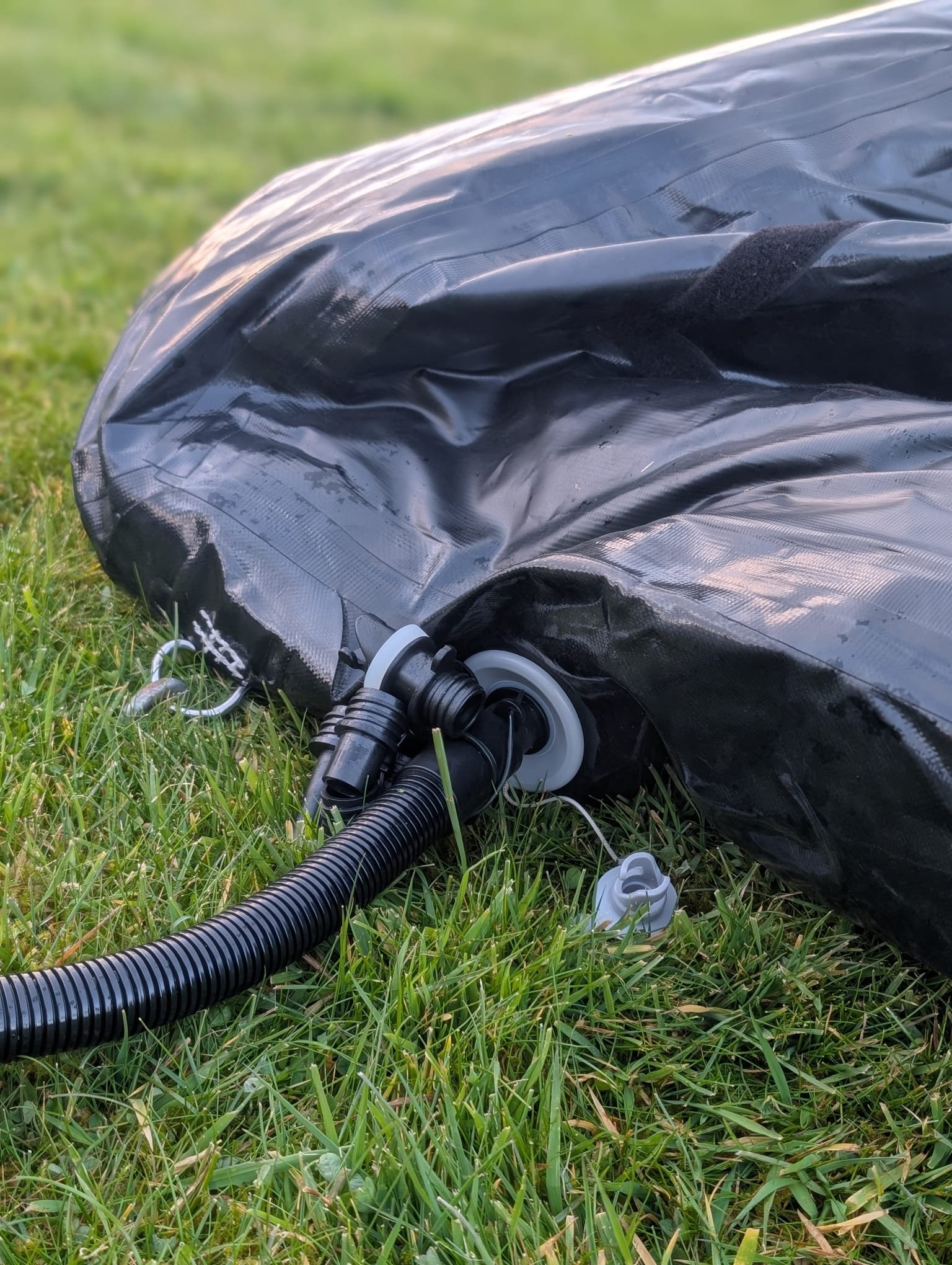 A black inflatable or portable item with a hose and white fittings attached, lying on green grass.