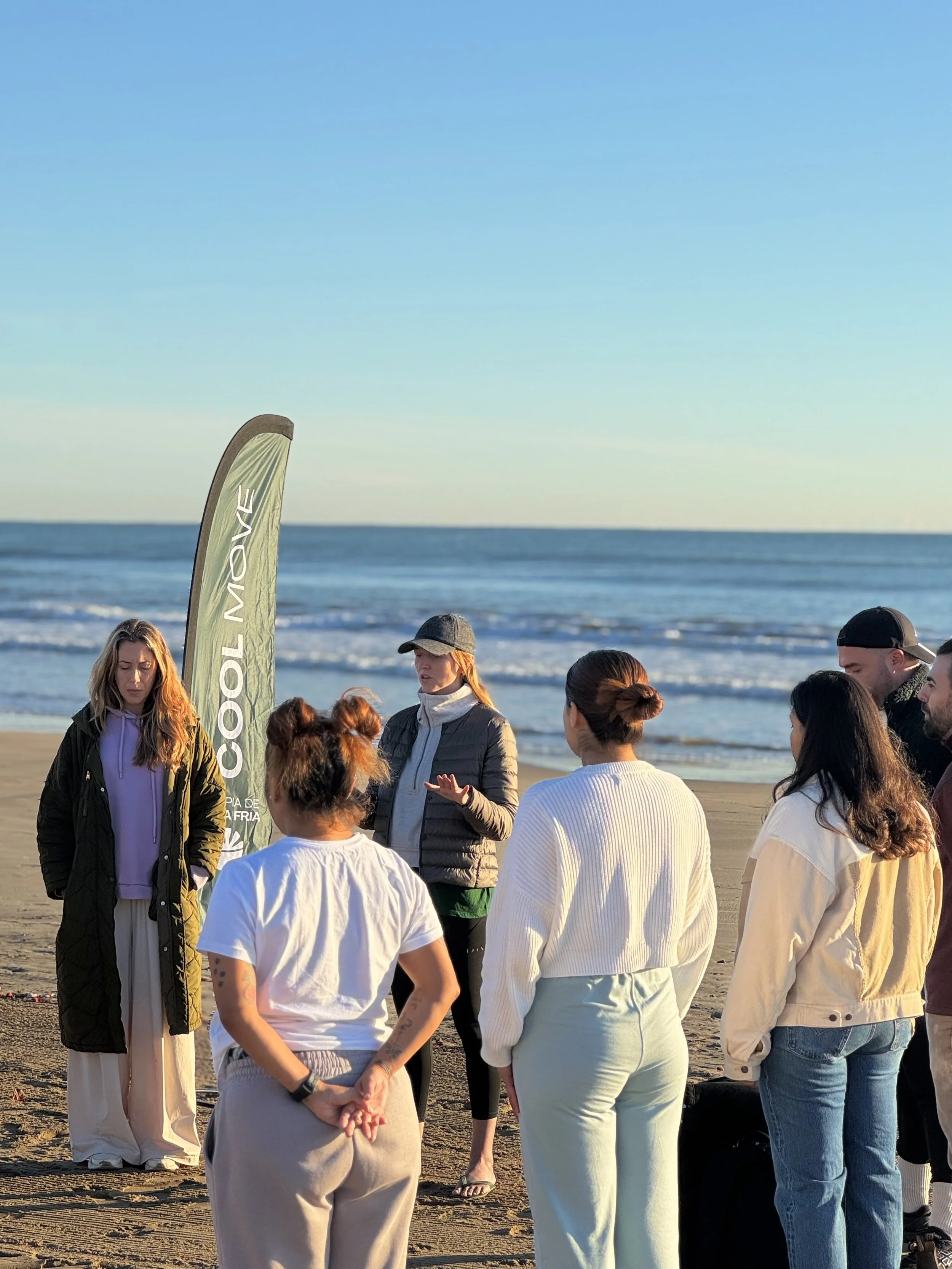 Grupo de personas reunidas en una playa, conversando cerca del mar. Una bandera dice "MOVE". Cielo despejado, ambiente soleado.