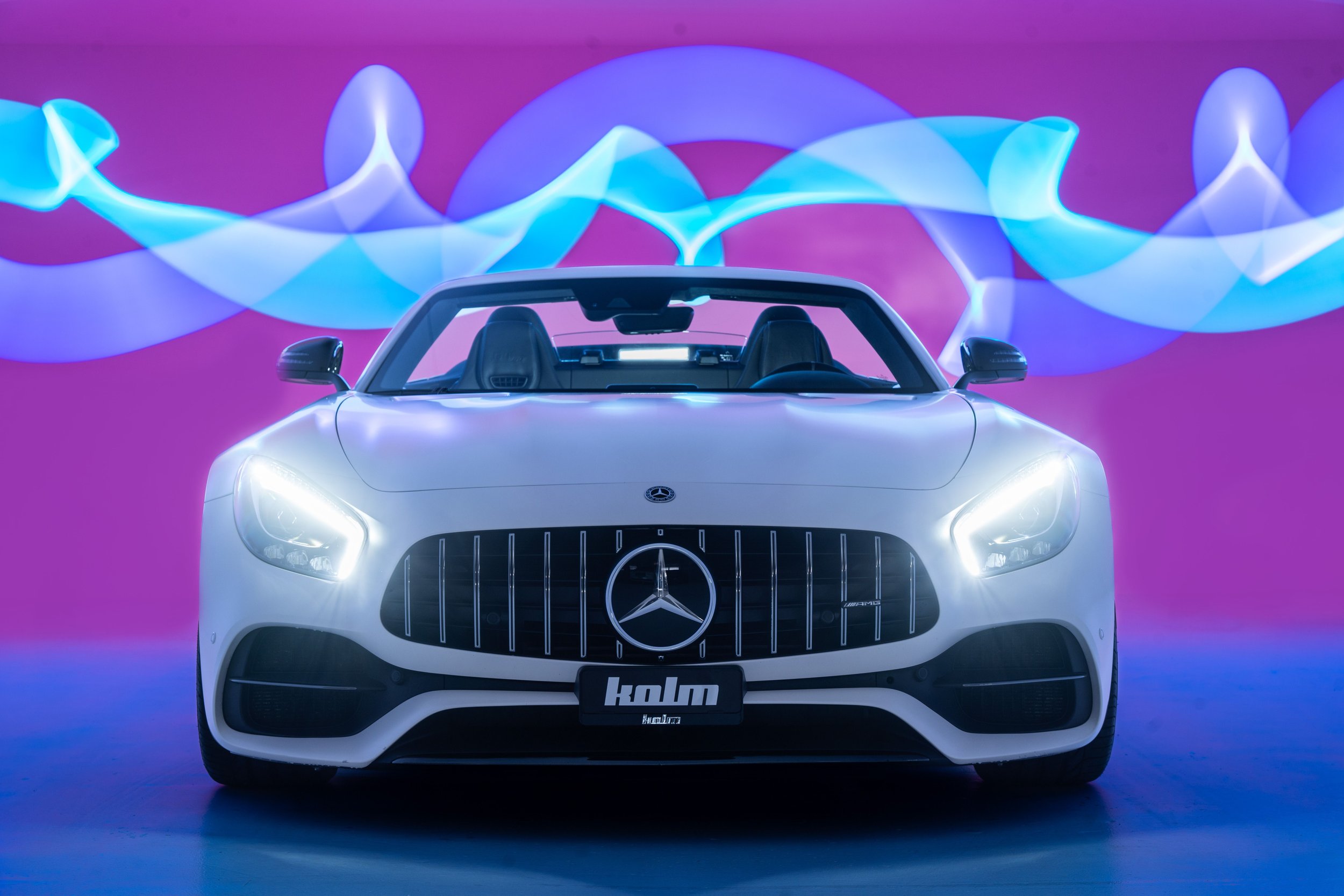 Ein weißer Sportwagen der Marke Mercedes-Benz steht frontal vor einem rosa Hintergrund mit blauen Lichtstreifen.
