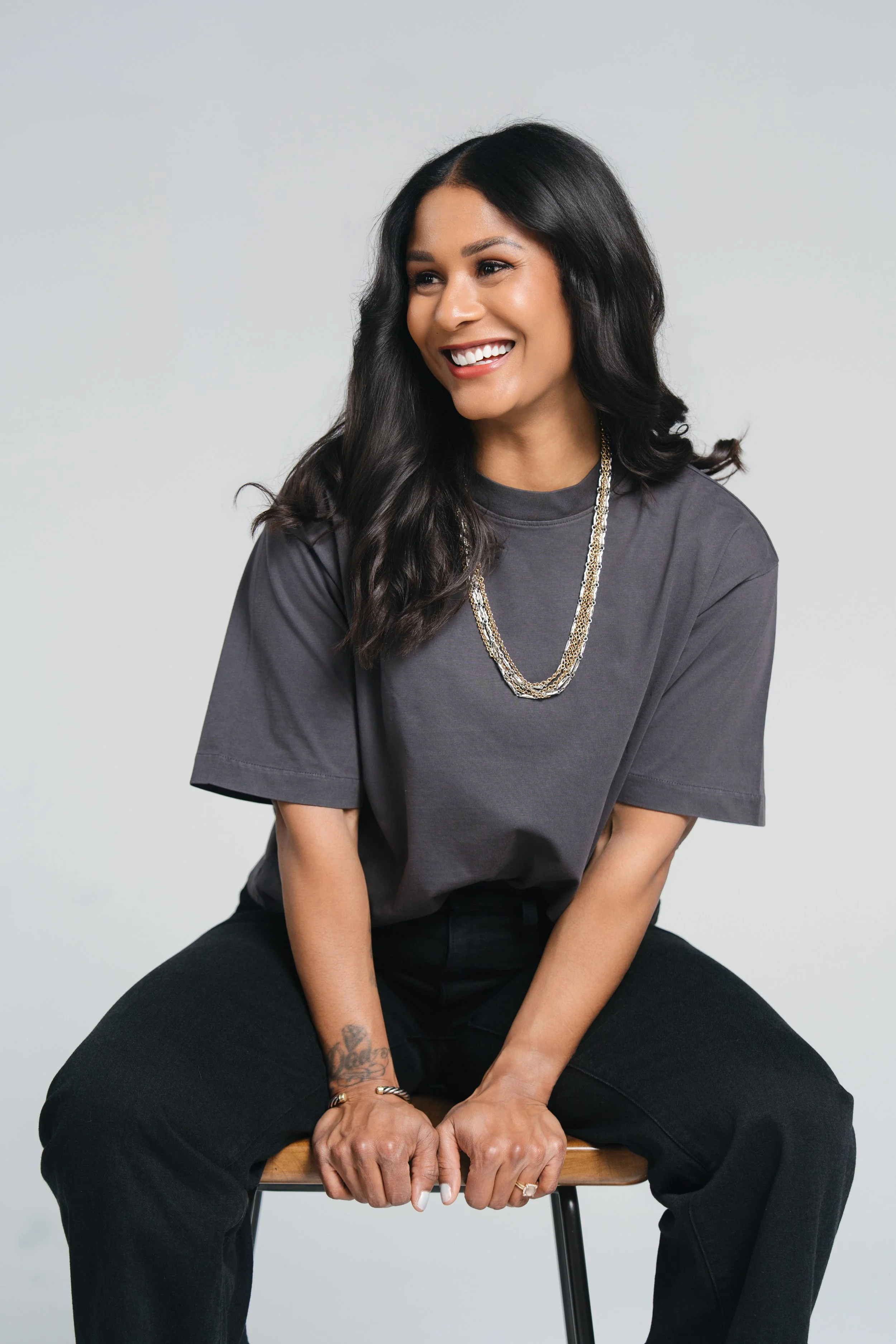 Woman with dark hair, wearing a gray t-shirt and jewelry, sitting on a stool against a plain background, smiling and looking to the side.