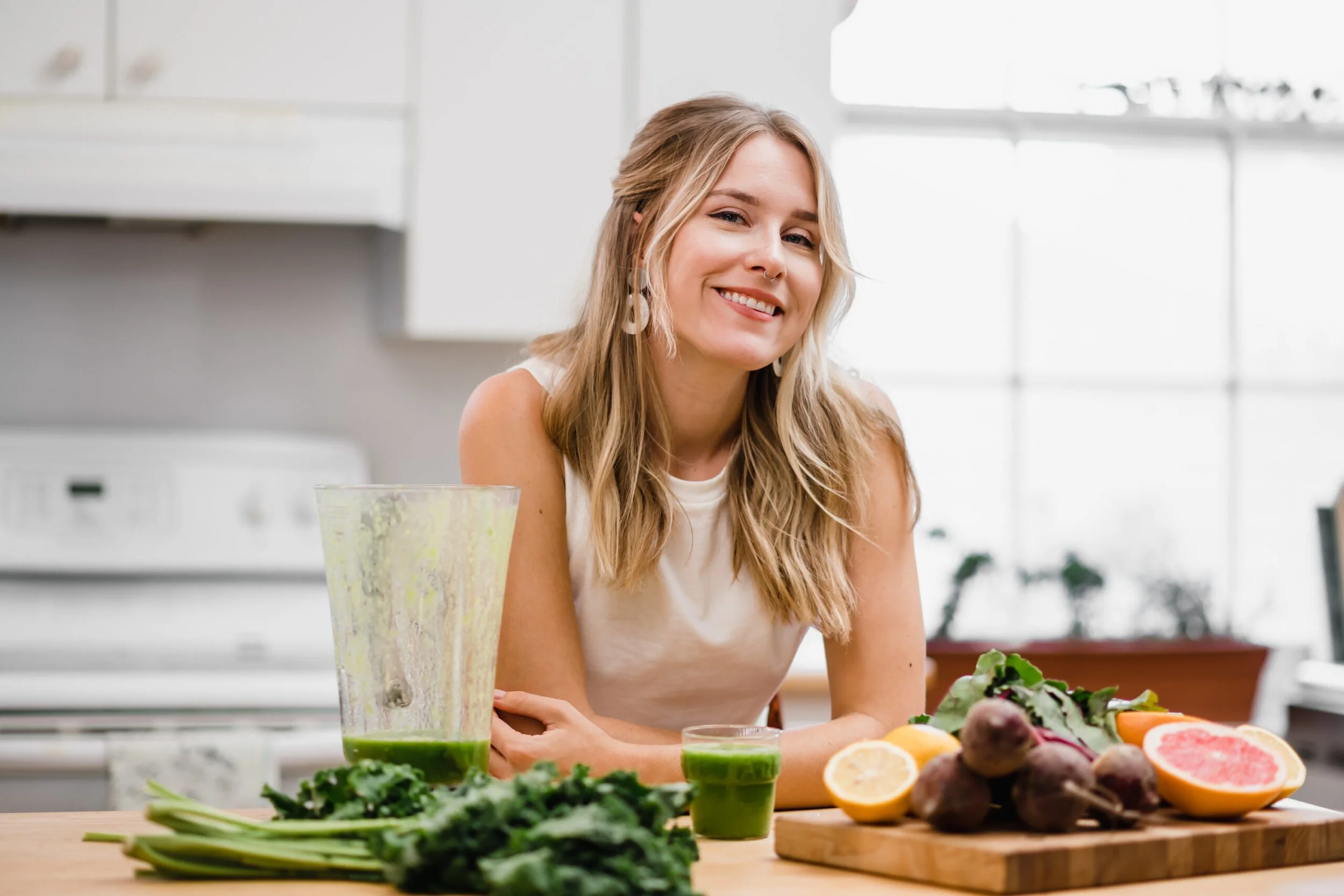 A woman with blonde hair and large earrings smiling at a kitchen counter with various vegetables and a large green smoothie in front of her.