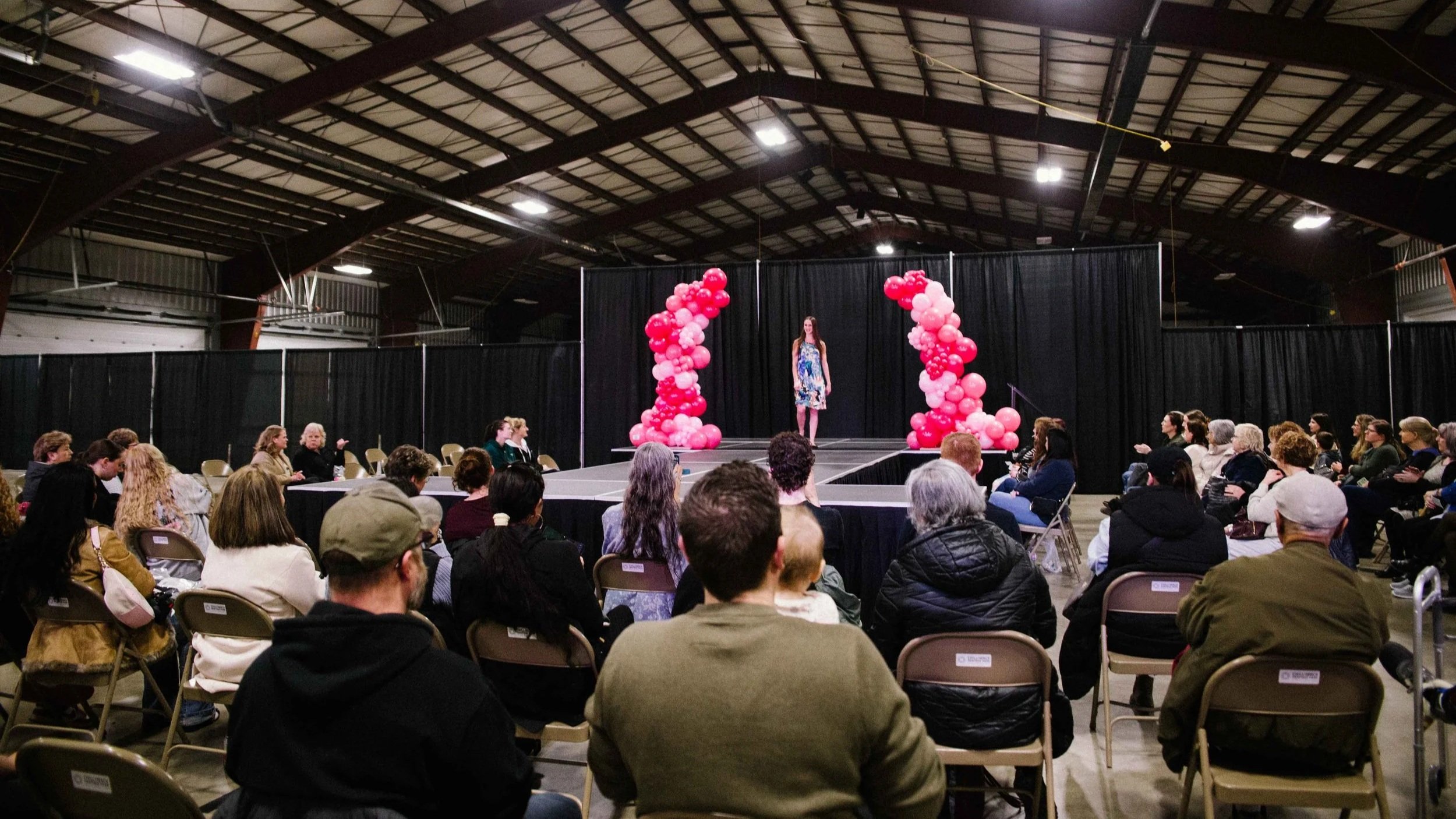 A woman standing on a stage decorated with pink balloons, addressing an audience seated in rows inside a large indoor venue with a high ceiling.