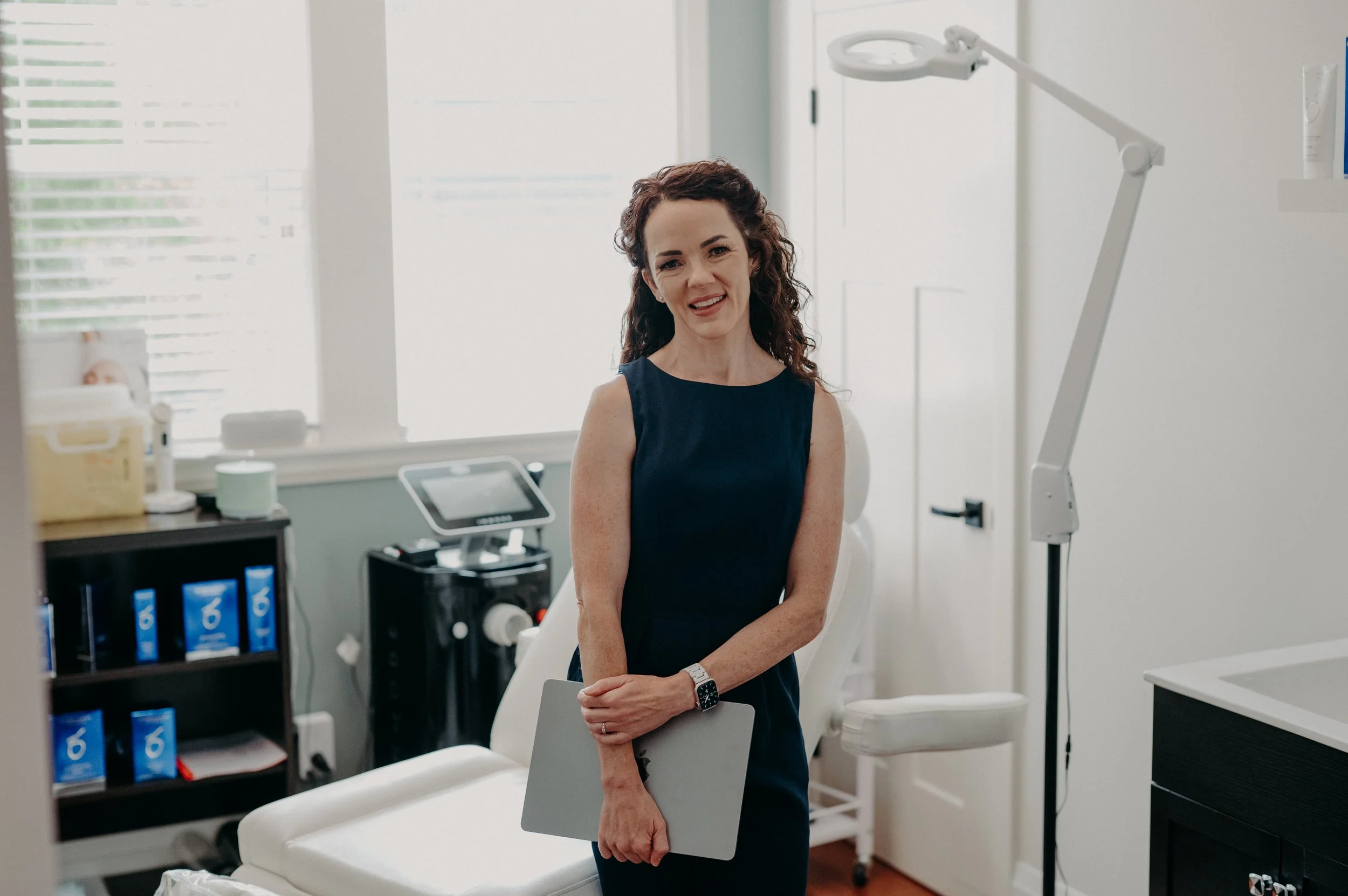 A woman with curly brown hair wearing a dark sleeveless dress and a watch on her left wrist, holding a clipboard, standing in a medical or clinical setting.