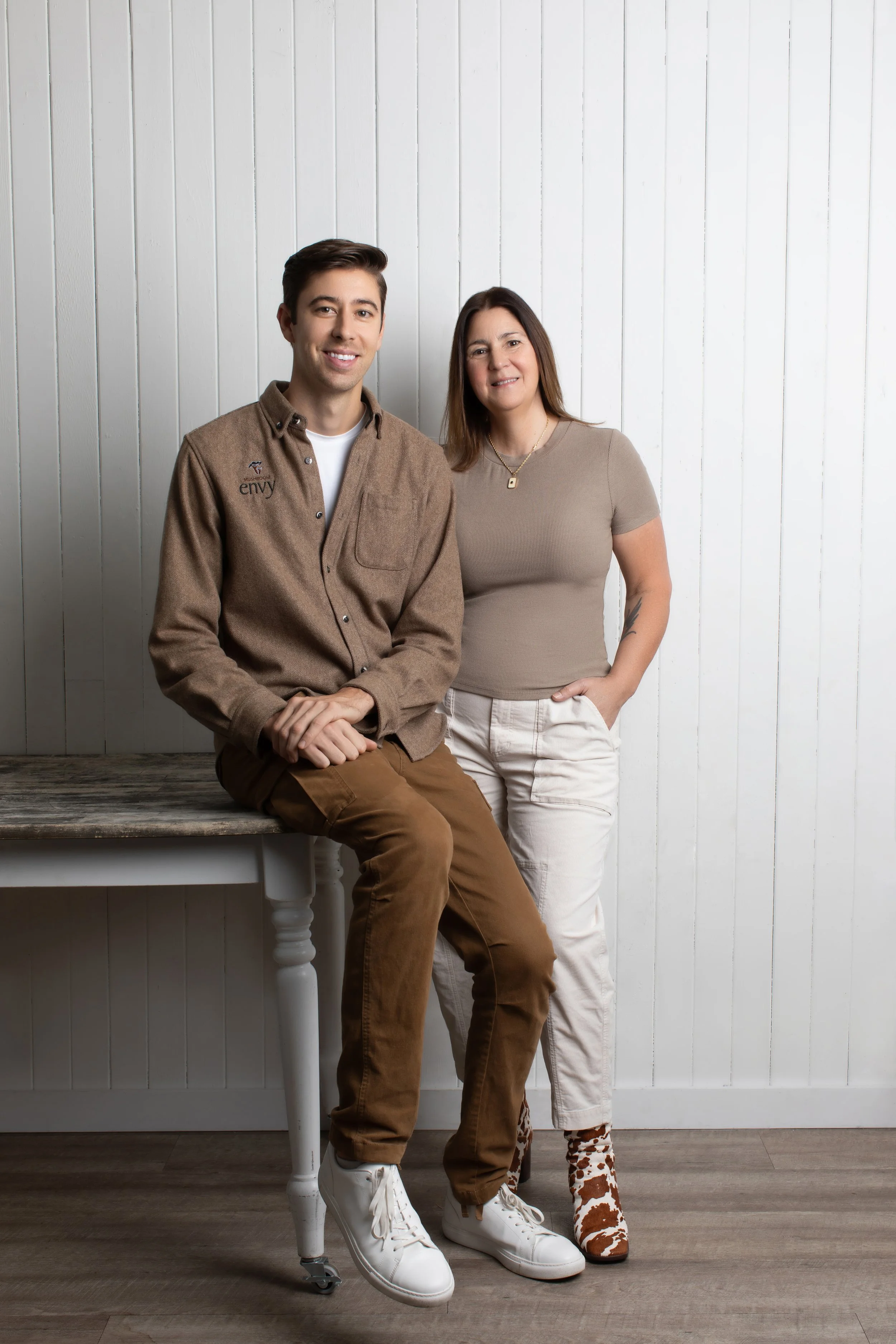 A young man sitting on a white wooden table with a woman standing next to him, both smiling and facing the camera in a room with white paneled walls.