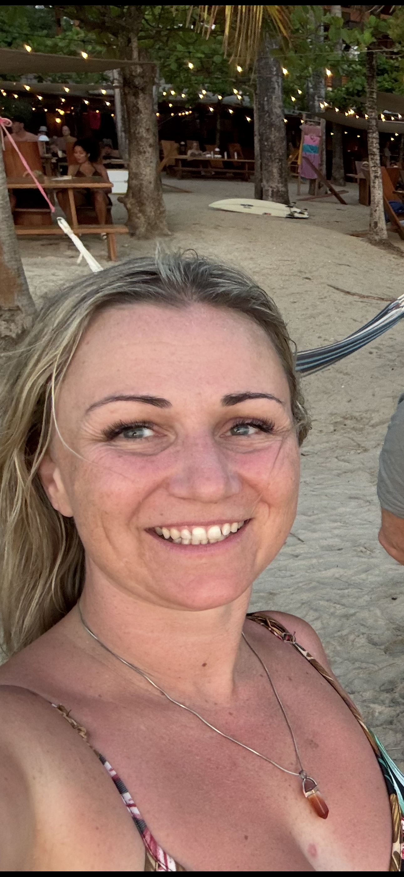 Woman smiling on the beach with sand, trees, and string lights in the background.