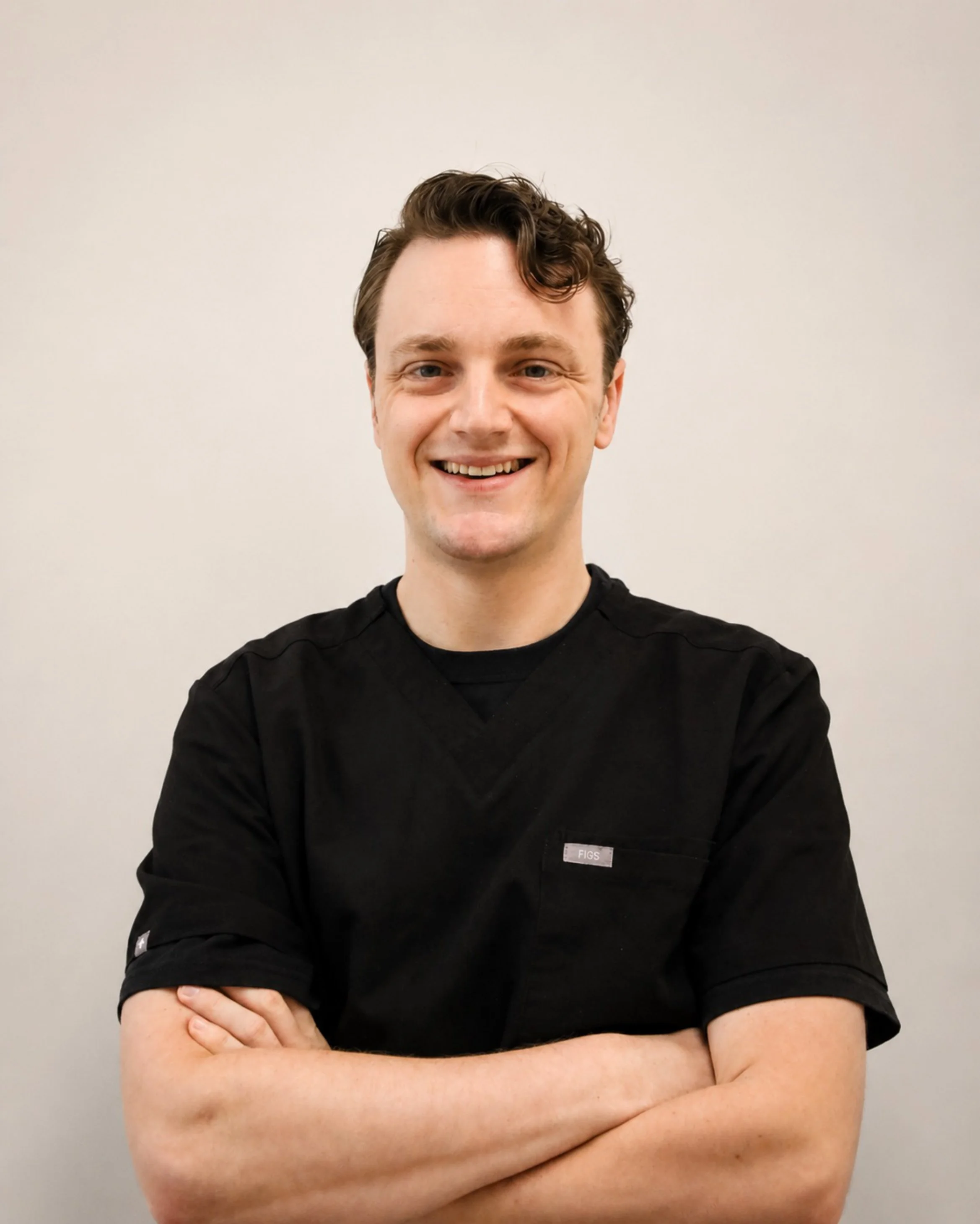 A young man with light skin and curly brown hair, smiling, standing against a plain white wall, wearing a black top with "FIGS" logo on the left sleeve, with arms crossed.