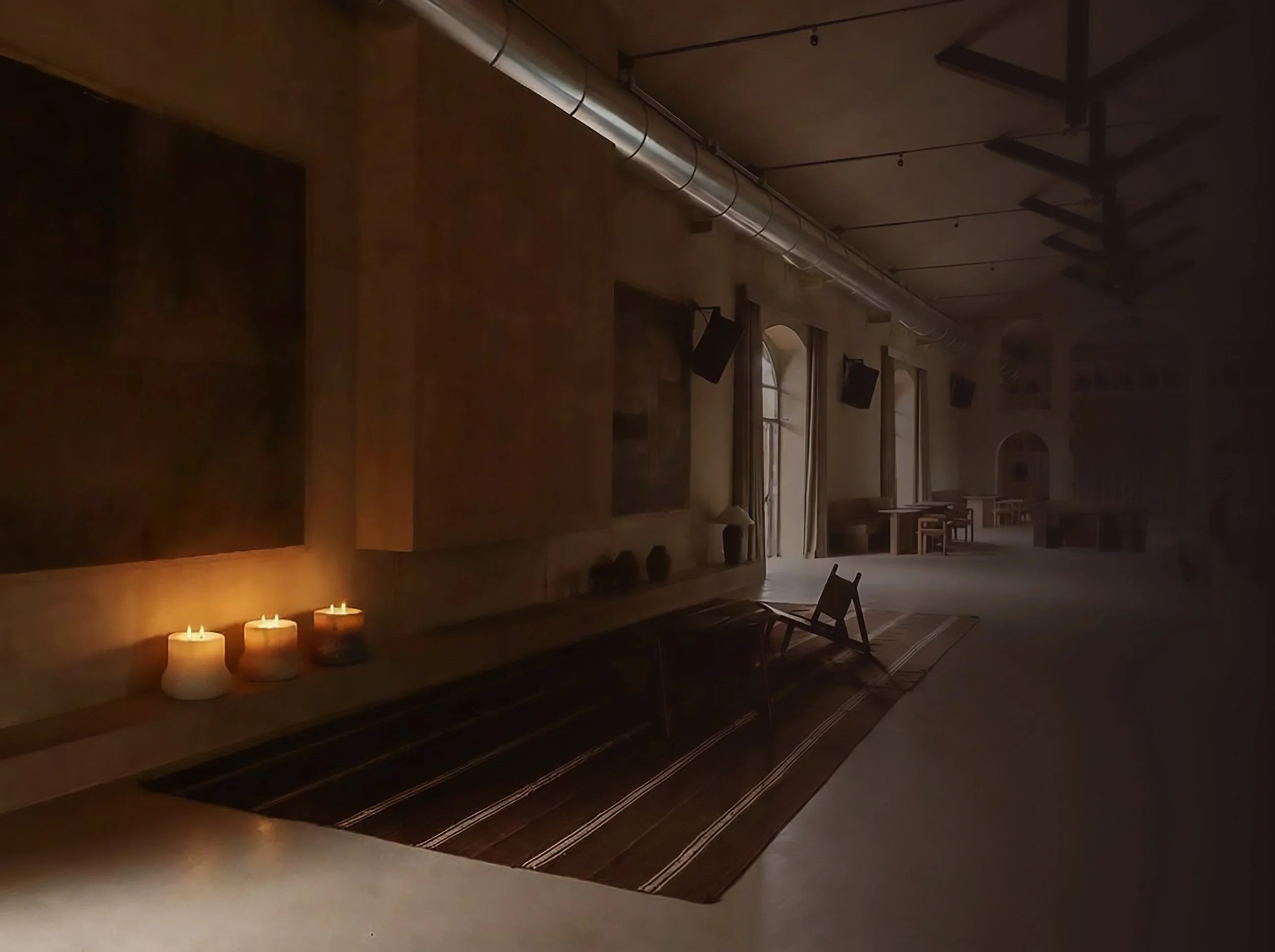 Dimly lit indoor space with candles, wooden chairs, and long patterned rugs on the floor. The room has large windows and a high ceiling with exposed beams and piping.