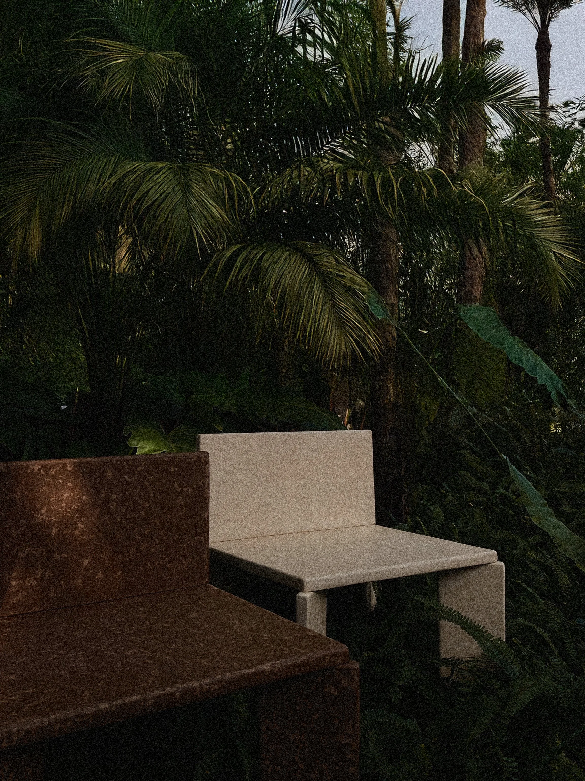 A stone bench with beige seating and reddish-brown armrests is situated outdoors among lush green tropical plants and tall palm trees.