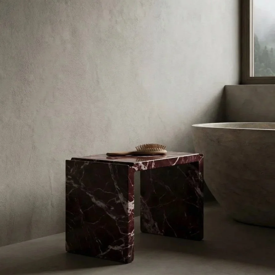 A small, rectangular dark red marble side table with white veins, holding a wooden hairbrush, next to a freestanding bathtub by a window.