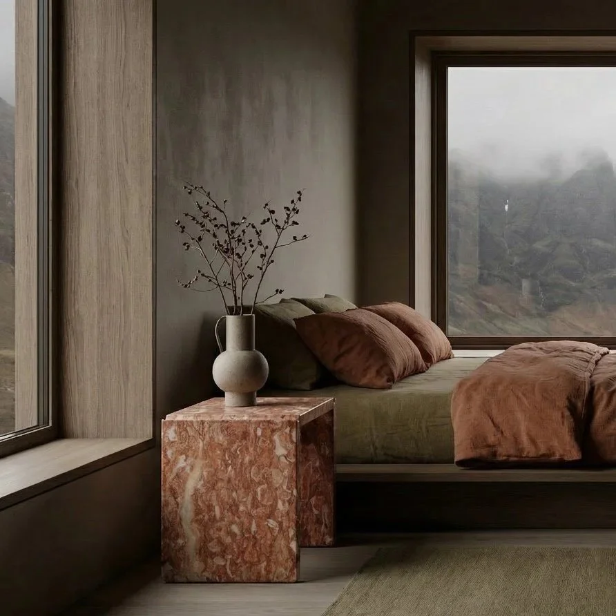 A cozy bedroom corner with a bed against a wall, dark pillows, and rust-colored bedding. Next to the bed is a small marble side table with a tall, narrow vase holding dried branches. Large windows reveal a misty mountainous landscape outside.