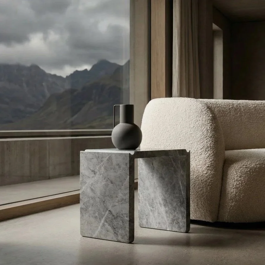 Interior of a modern room with a large window showing mountains and stormy skies outside, a beige textured sofa, and a small marble side table with a black vase.