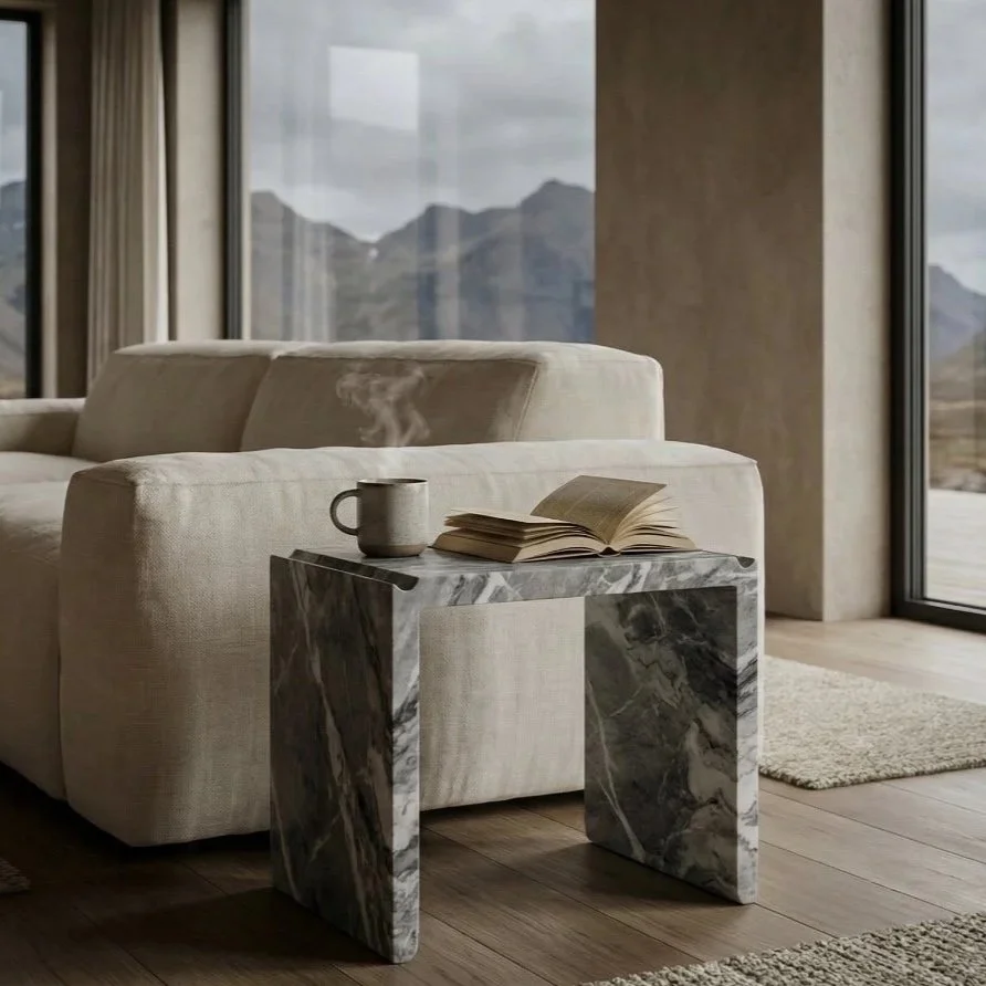 A living room with a beige sofa, marble side table with coffee mug and open book, large windows showing mountain landscape, wooden floor, and neutral decor.