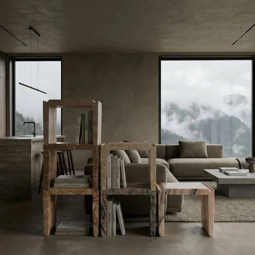 A modern living room with large windows showing foggy mountain views, a beige sofa with throw pillows, a marble coffee table, a bookshelf with books, and a minimalist kitchen area with a sink and faucet.