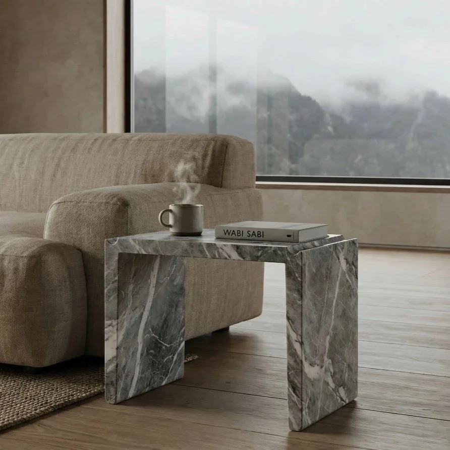 A beige sofa next to a marble side table with a steaming mug and a book titled 'WABI SABI' on it, in front of a large window with a mountain view.