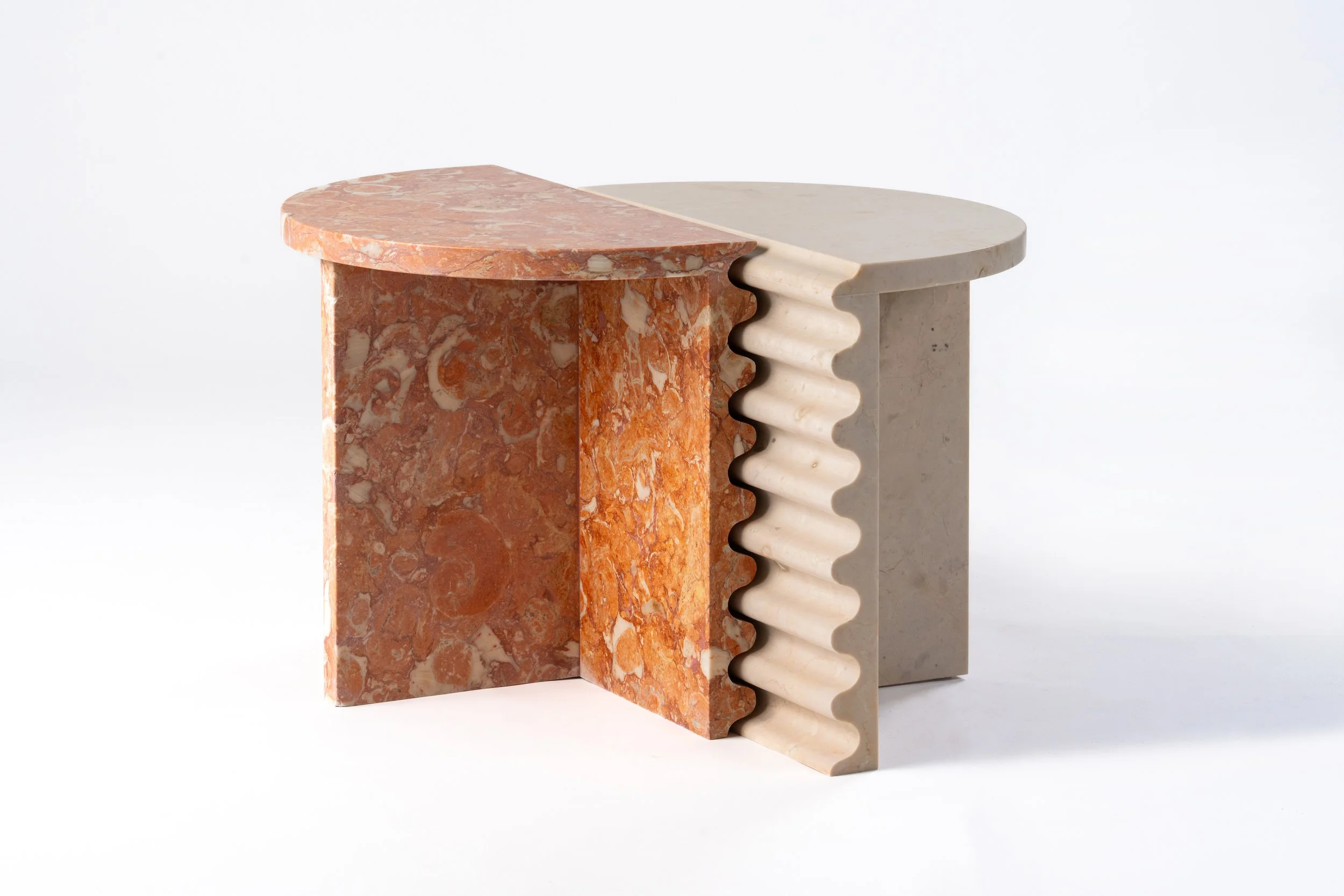 A modern sculpture made of three vertically aligned marble blocks with different textures and colors. The left block is pinkish-red with a marbled pattern, the middle is a pinkish-orange with a similar pattern, and the right block is off-white with a smooth, ridged texture. All three blocks are supported on a flat base and arranged against a plain white background.