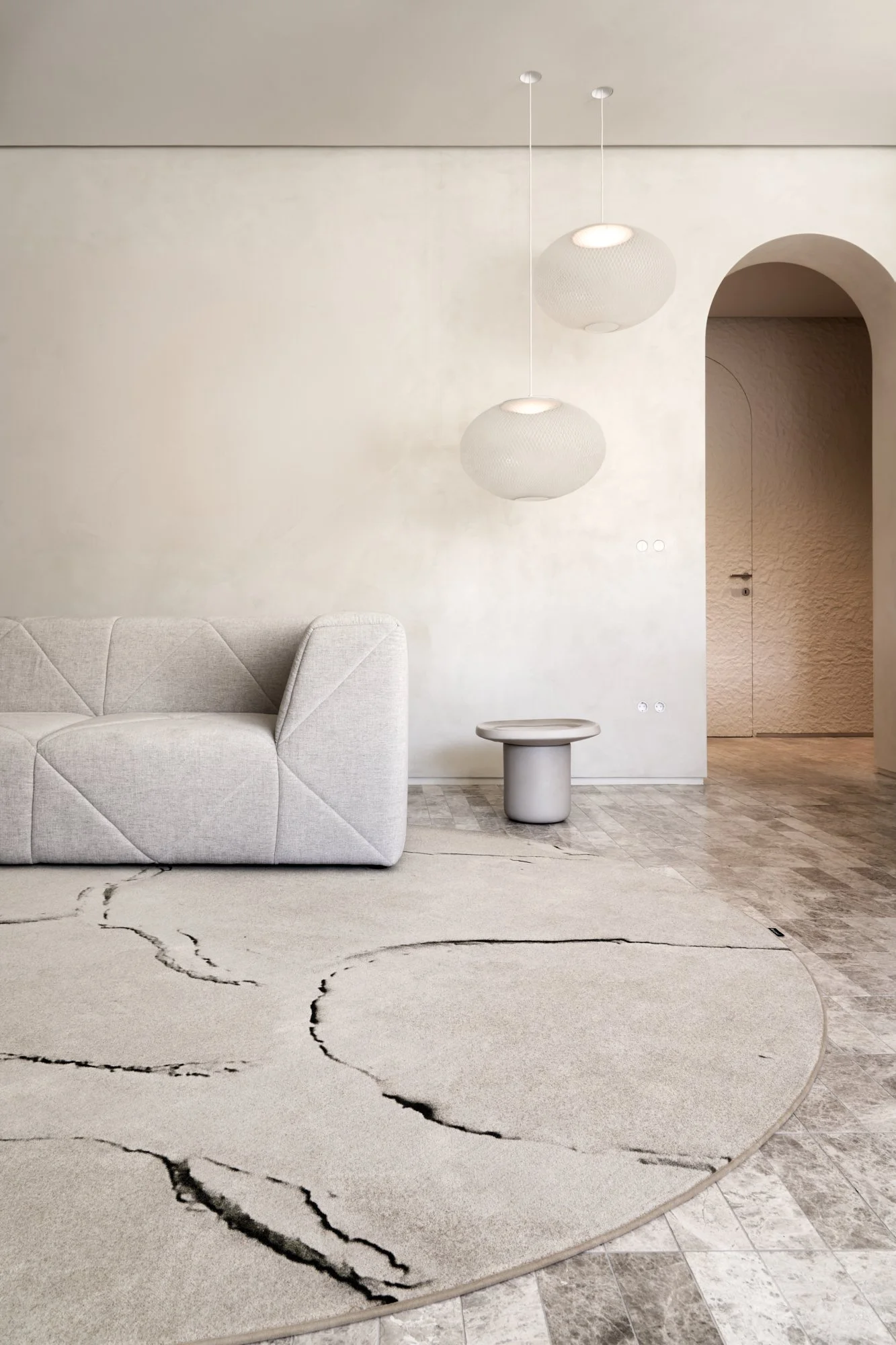 Minimalist living room with a light-colored sofa, a round side table, and two modern hanging pendant lights. There is a decorative area rug with black marbled accents on a tile floor.
