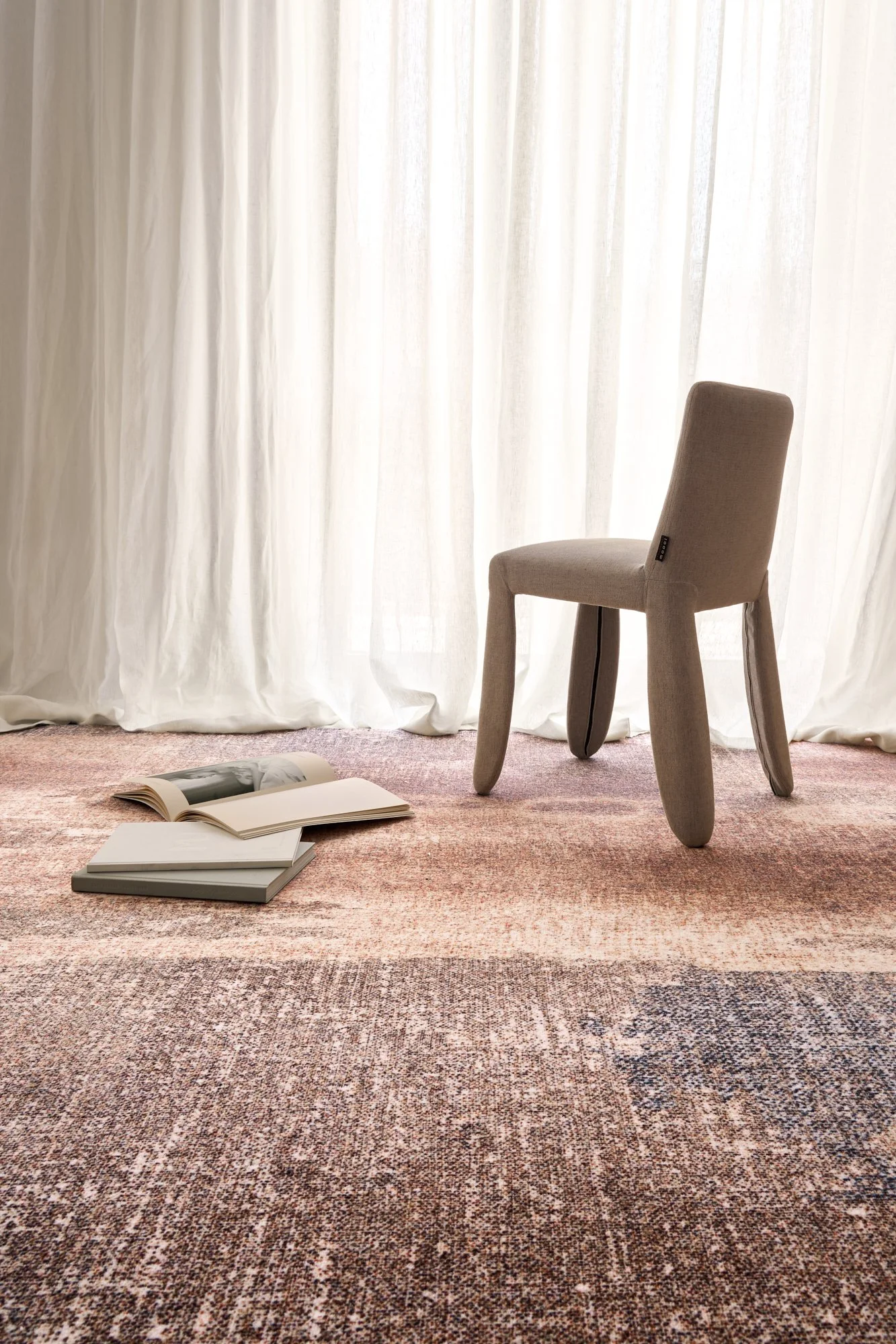 A beige upholstered chair sits on a multicolored textured rug in front of white curtains, with an open magazine and two closed books on the floor.