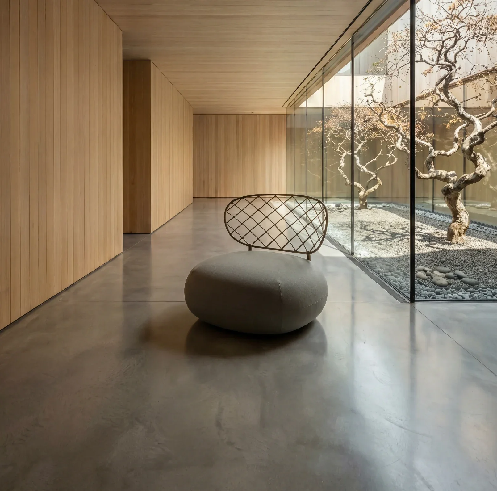 Modern interior space with light wood paneling on walls and ceiling, polished concrete floor, and a large glass window showing a courtyard with twisted trees and pebbles. A single rounded lounge chair with a lattice design on the backrest is in the foreground.