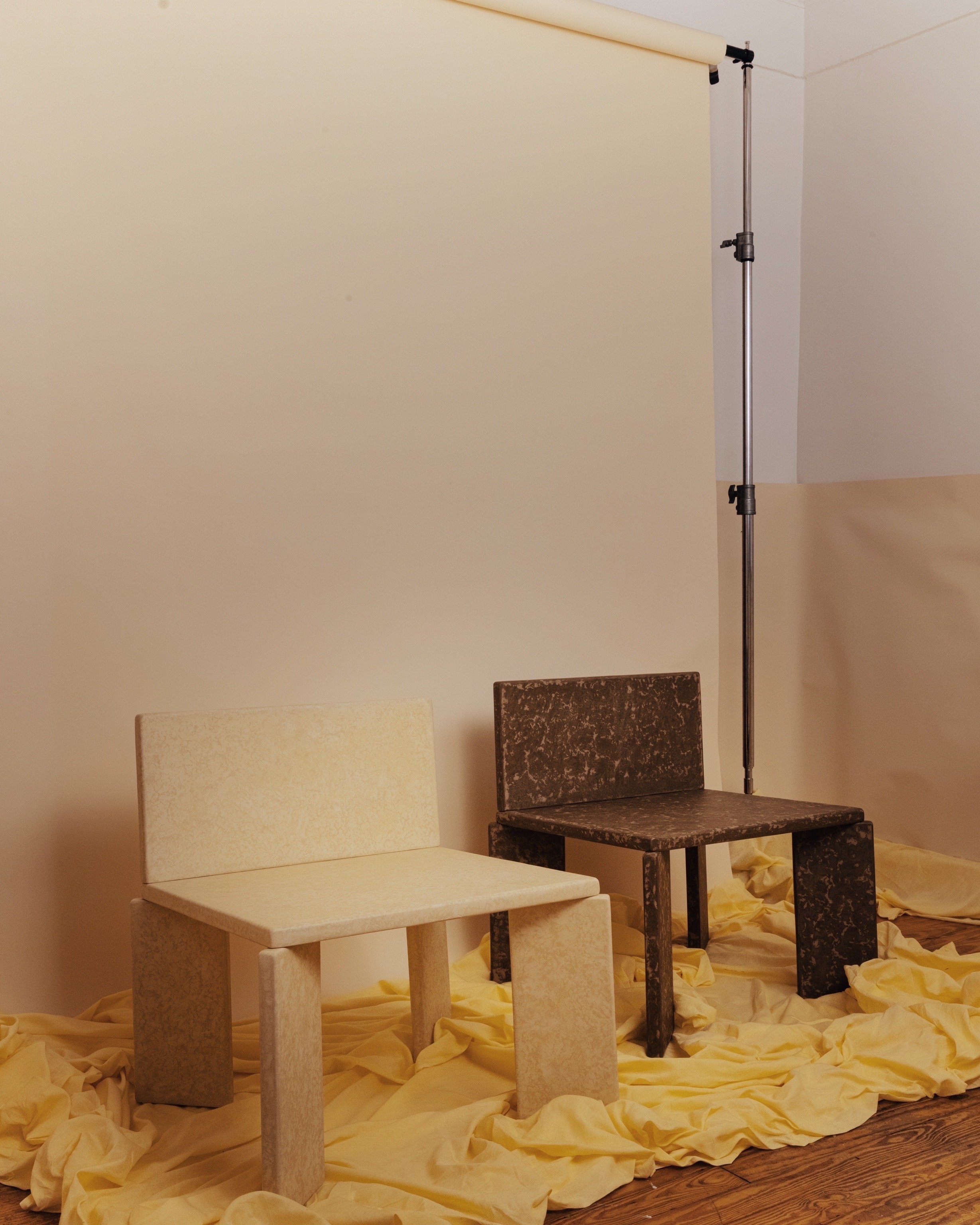 Display of two chairs, one white and one black, placed on a yellow fabric backdrop with a beige wall and a partial view of a wooden floor. There is also a white backdrop stand on the right side of the chairs.