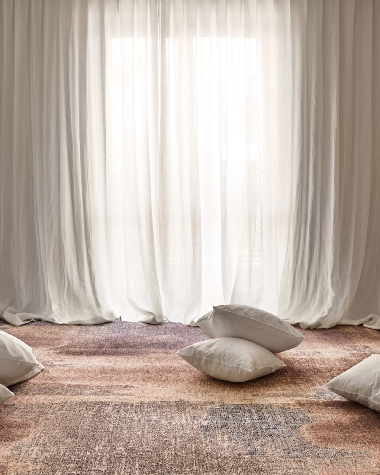 Cream-colored curtains hanging in front of a window, with several pillows on a patterned area rug on the floor.