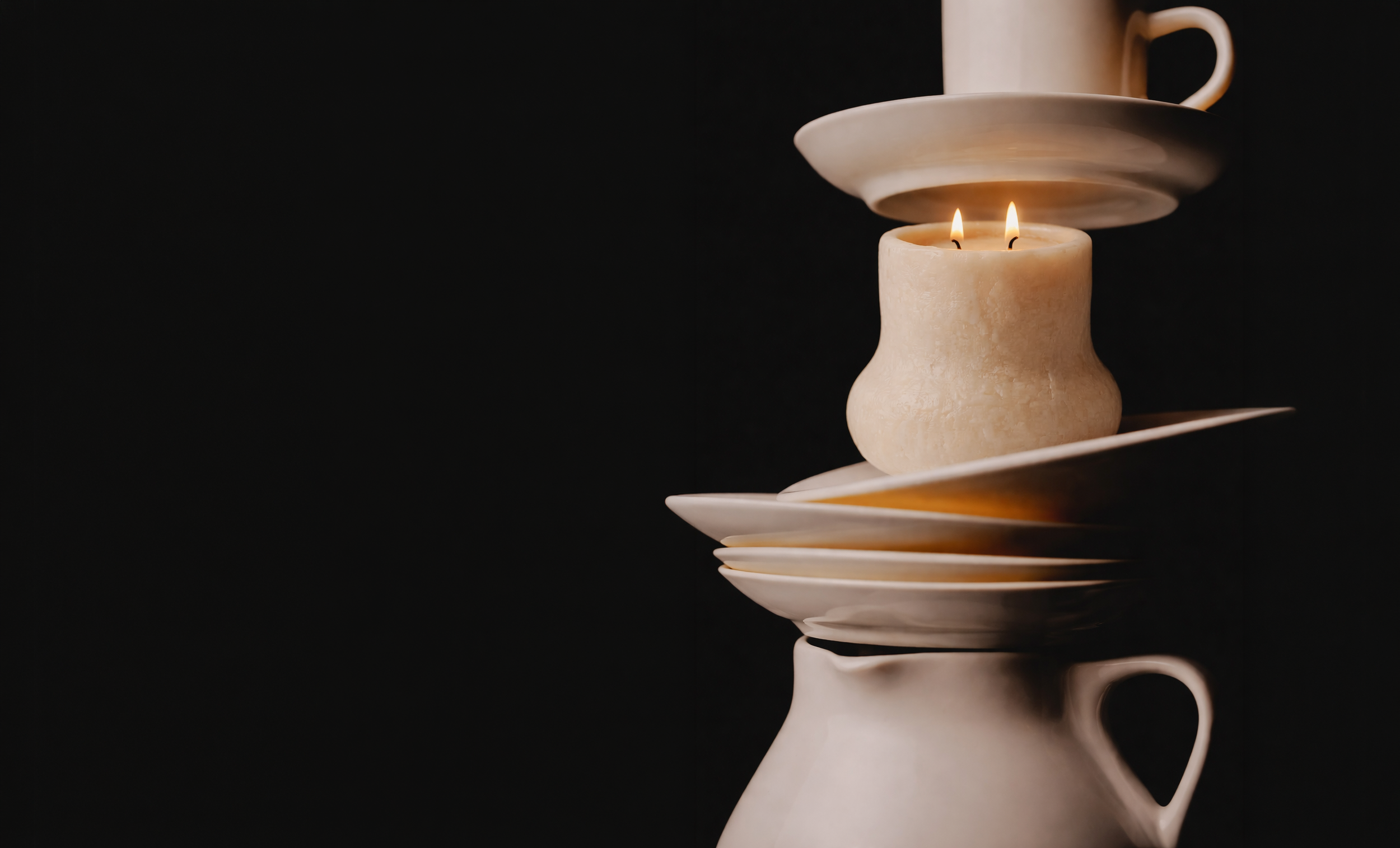 A cream-colored candle with two wicks burning, placed on a white plate, with stacked white dishes and a cup in the background, set against a black backdrop.
