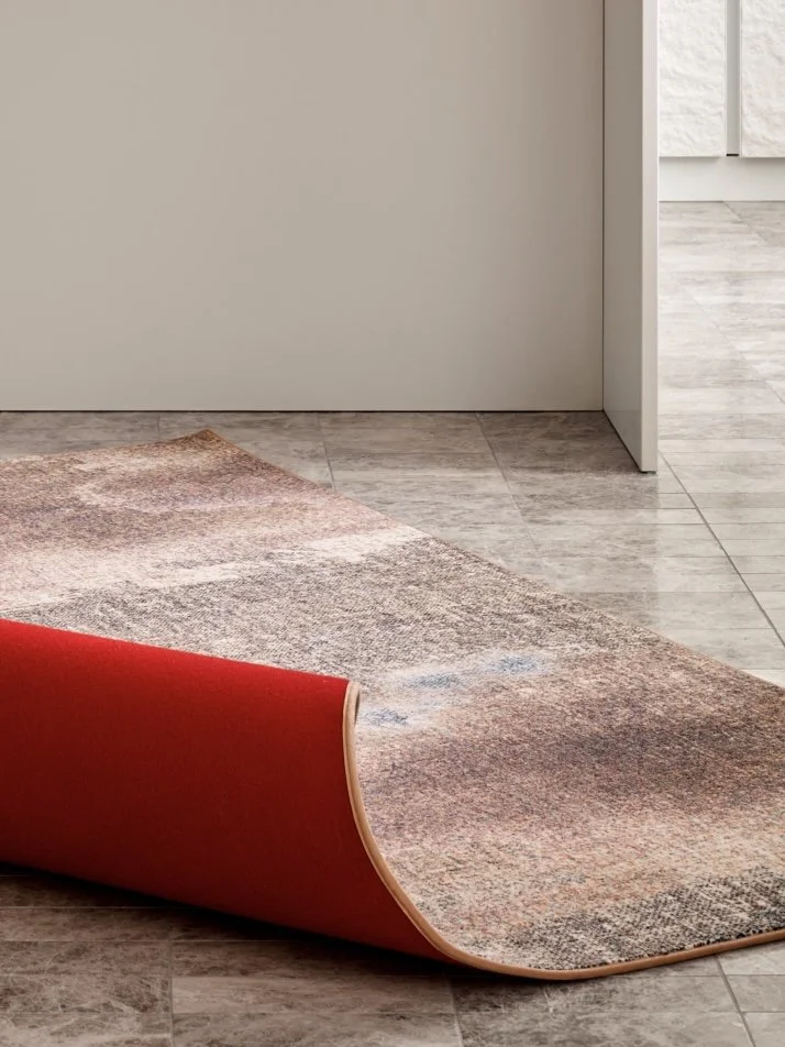 Part of a multicolored area rug with a red felt backing, partially rolled up on a wood and tile floor in a neutral-colored room.
