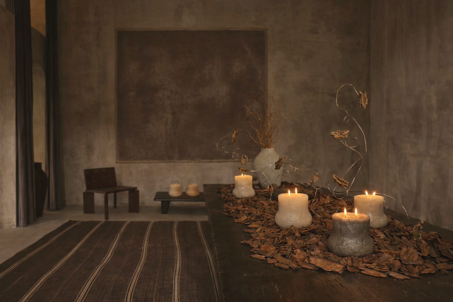 A room with a table decorated with lit candles, dried leaves, and a decorative vase with dried branches. There are two chairs, a textured wall with a large empty frame or canvas, and a striped rug.