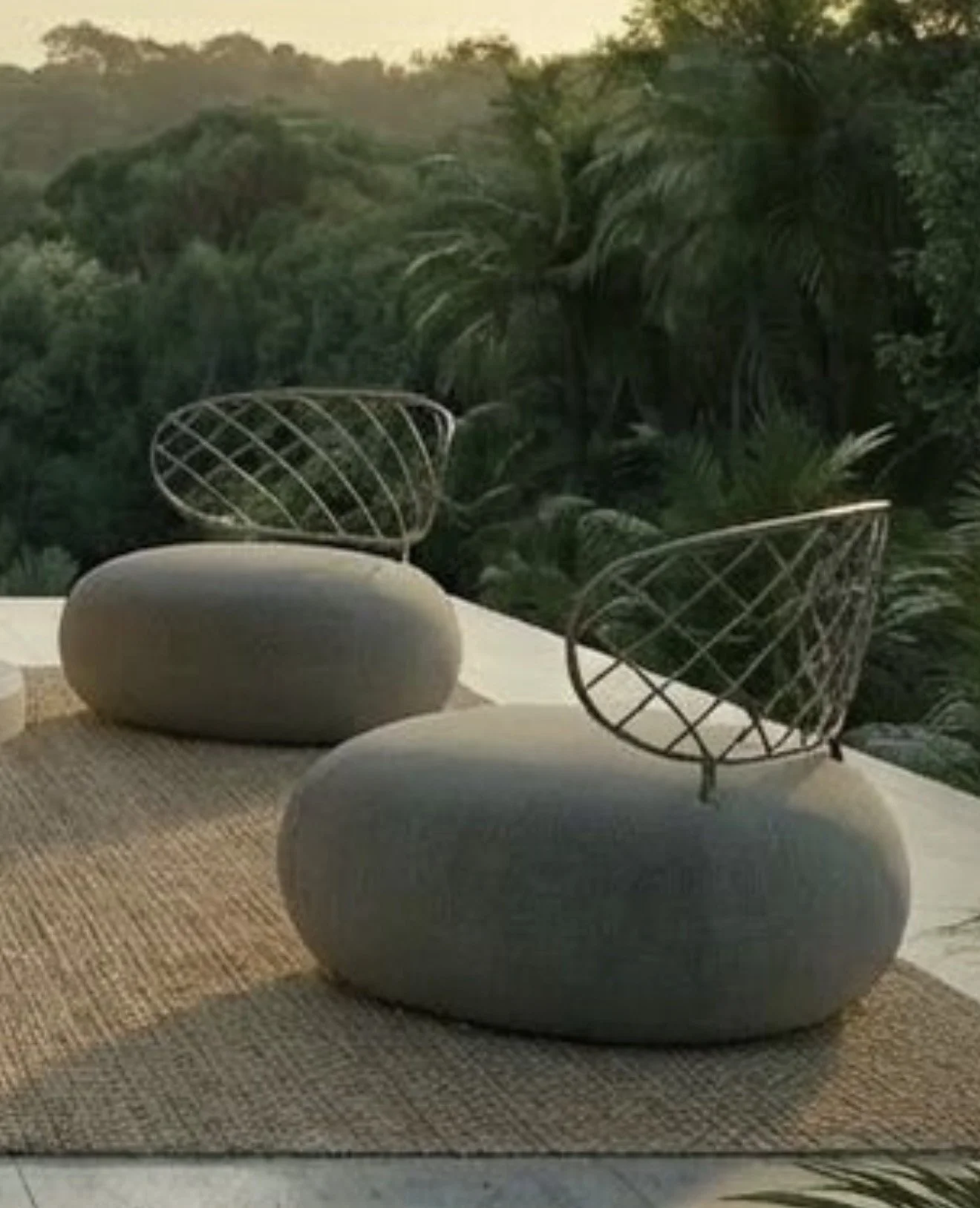Two modern wireframe chairs with rounded stone bases on a outdoor patio with a textured rug, overlooking lush green foliage and trees at sunset.
