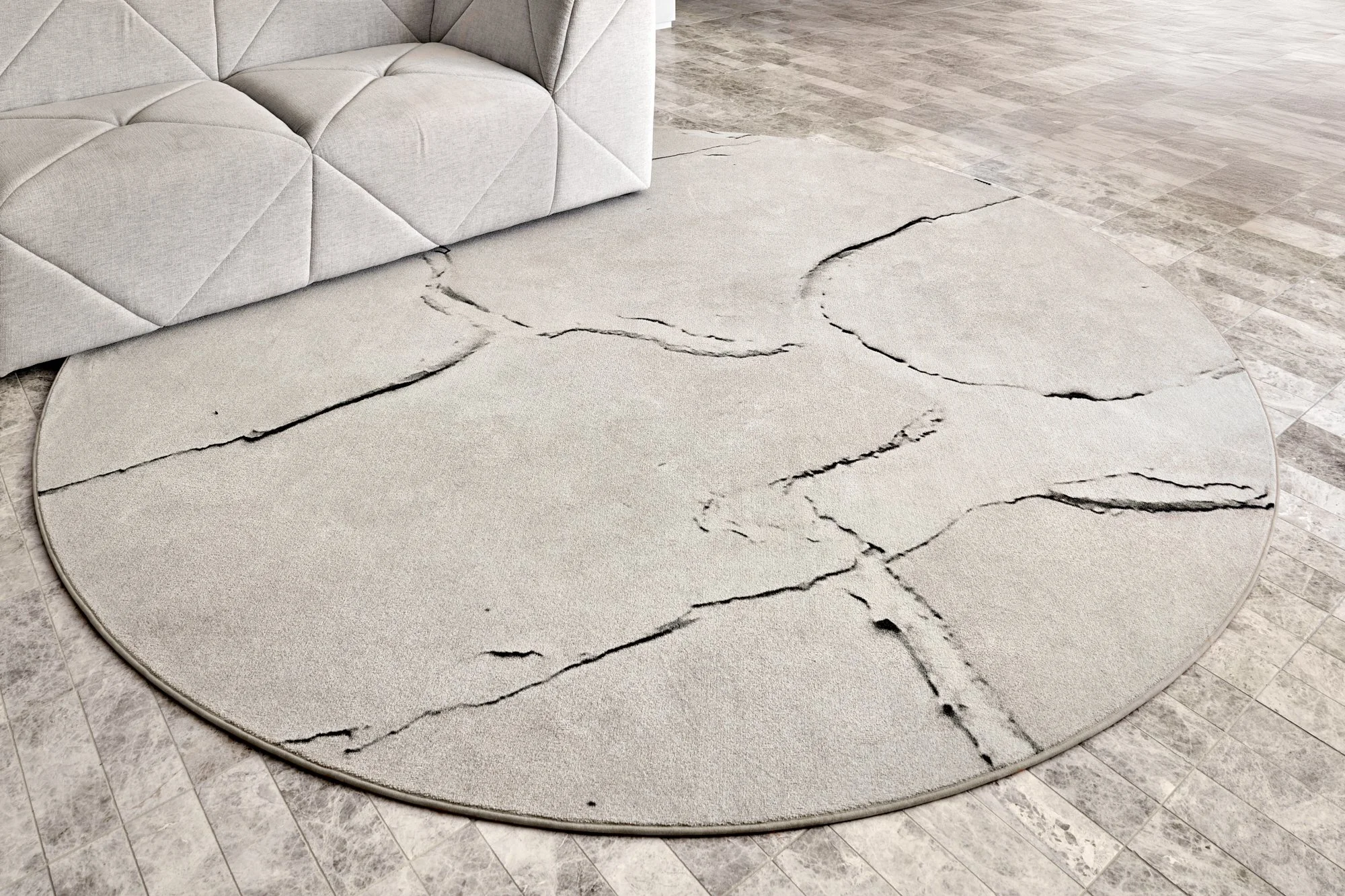 A round light-colored area rug with black cracks, placed on a tiled floor next to a light gray upholstered sofa that is slightly damaged with a tear on one corner.