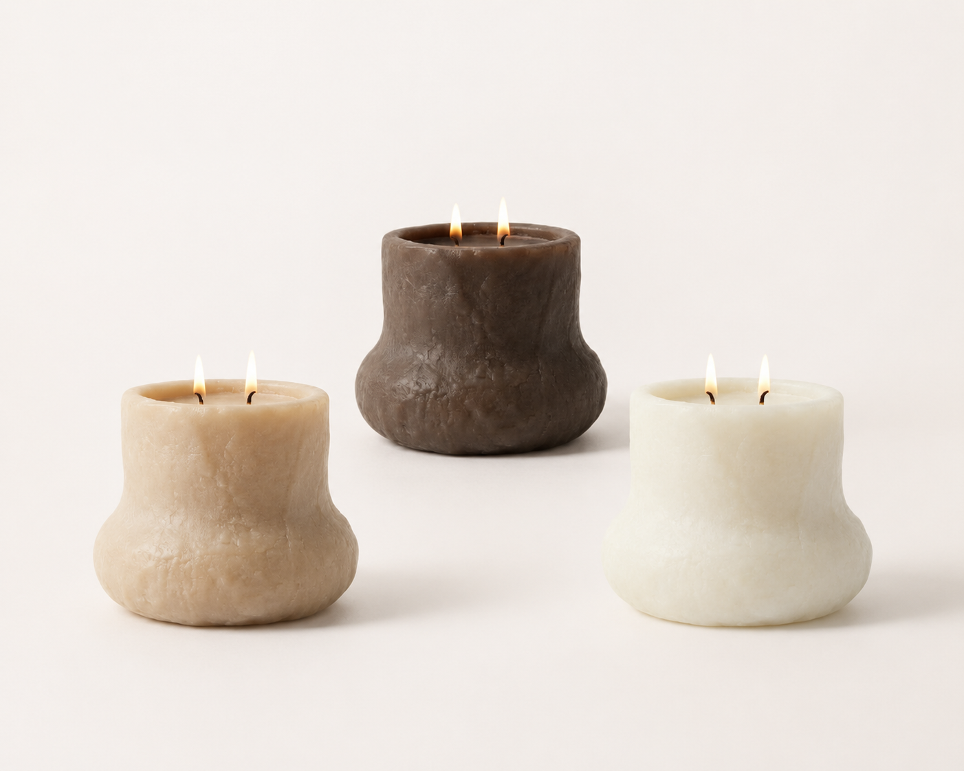 Three pillar candles with lit wicks, one cream-colored, one brown, and one white, placed on a plain white background.