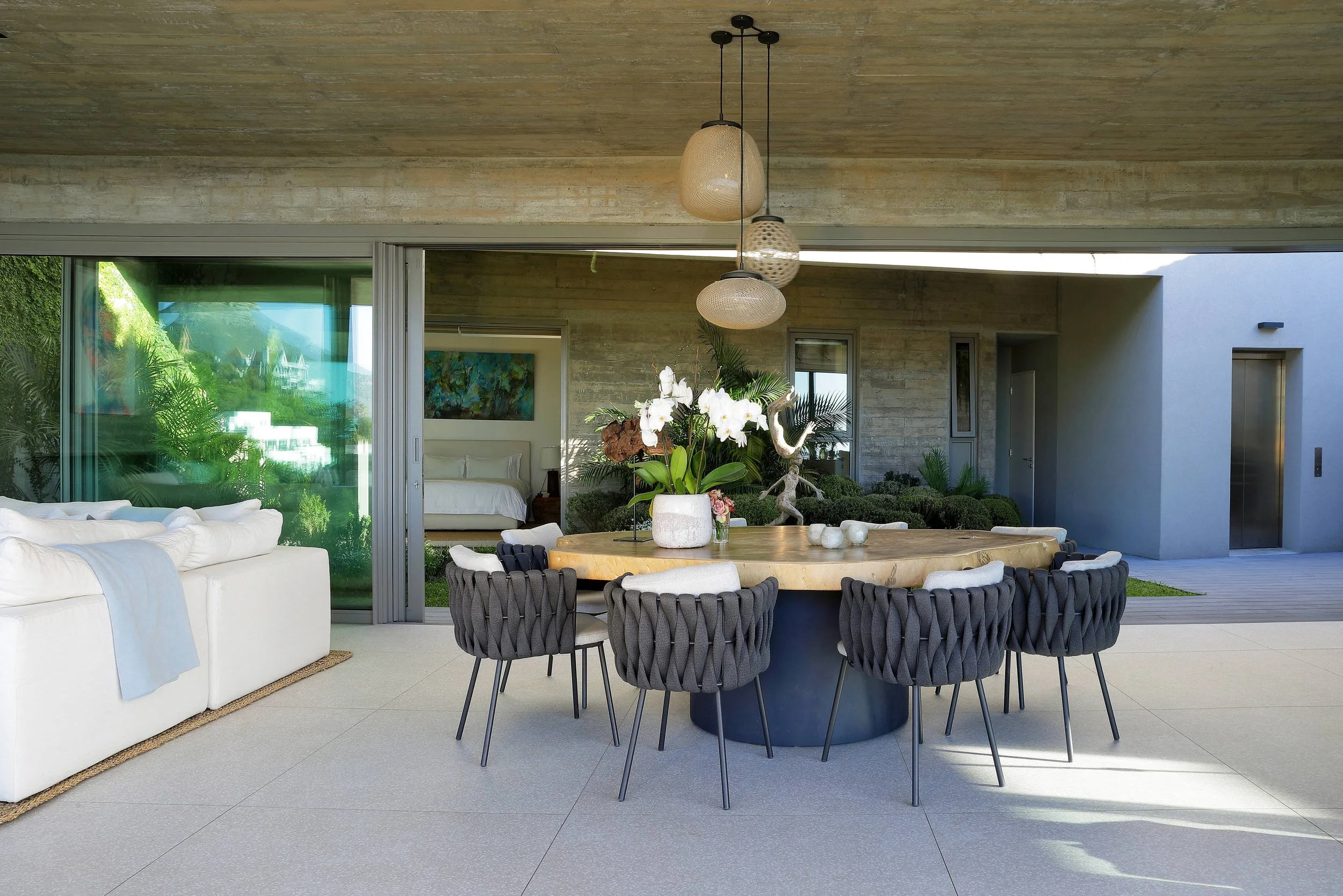 Moderner Essbereich im offenen Wohnraum mit runder Holztisch, schwarzen loungigen Sitzstühlen, Blumen, Vorhängen, Glaswänden und Blick in den Garten