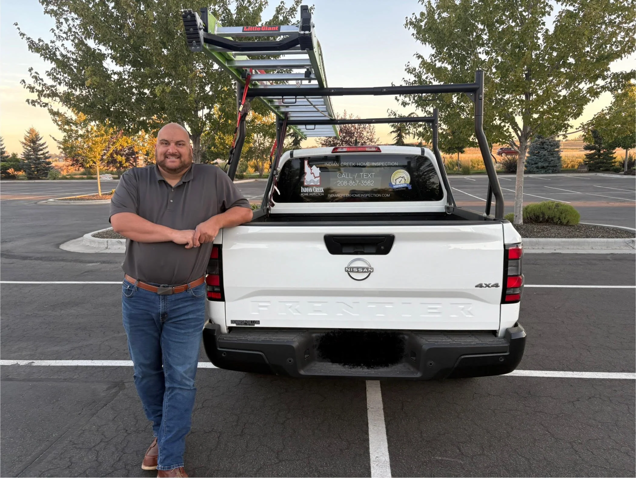 A man standing in a parking lot next to a white Nissan Frontier pickup truck with a ladder rack and promotional decals on the rear window for Indian Creek Home Inspection.