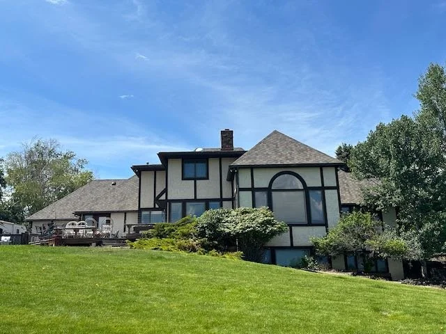 Large house with Tudor-style black and white exterior, surrounded by green lawn and trees, under a blue sky with clouds.