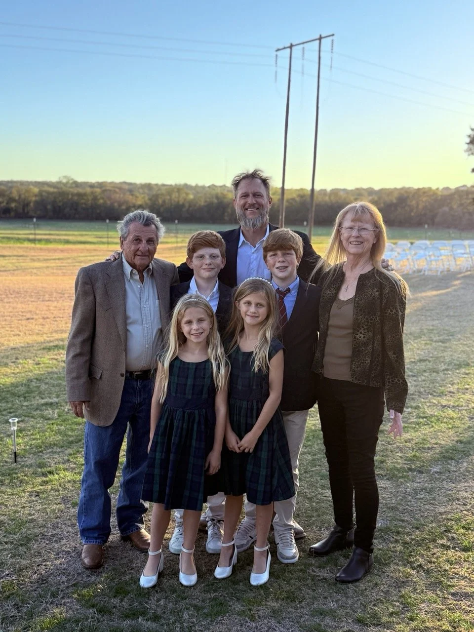 Family portrait of three generations outdoors during sunset, including two elderly adults, two middle-aged adults, two teenage boys, and two young girls, standing on grass with a field and trees in the background.