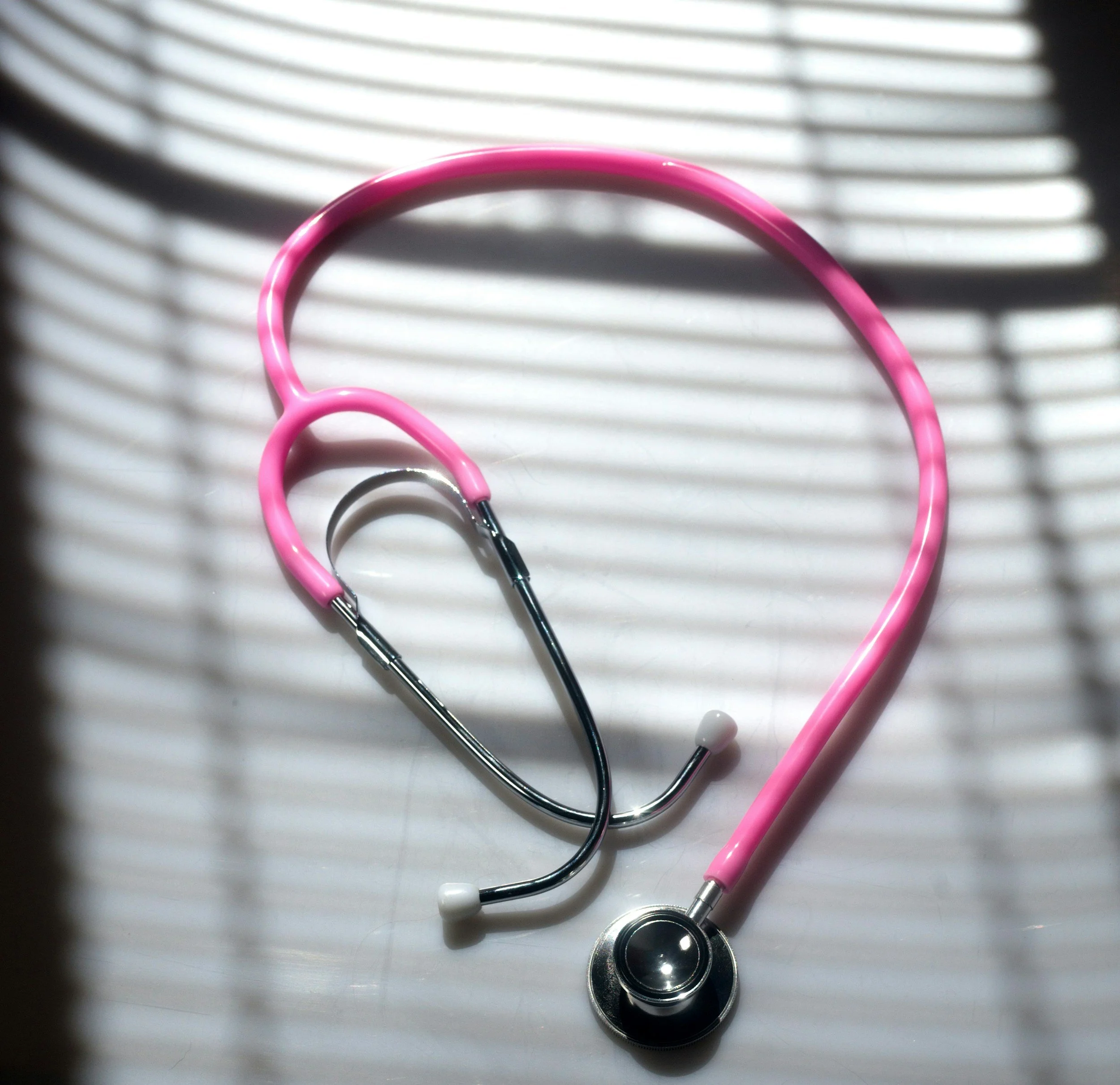 A close-up photograph of two stethoscopes lying on a surface with shadows cast in a striped pattern. One stethoscope has pink tubing and the other has black tubing.