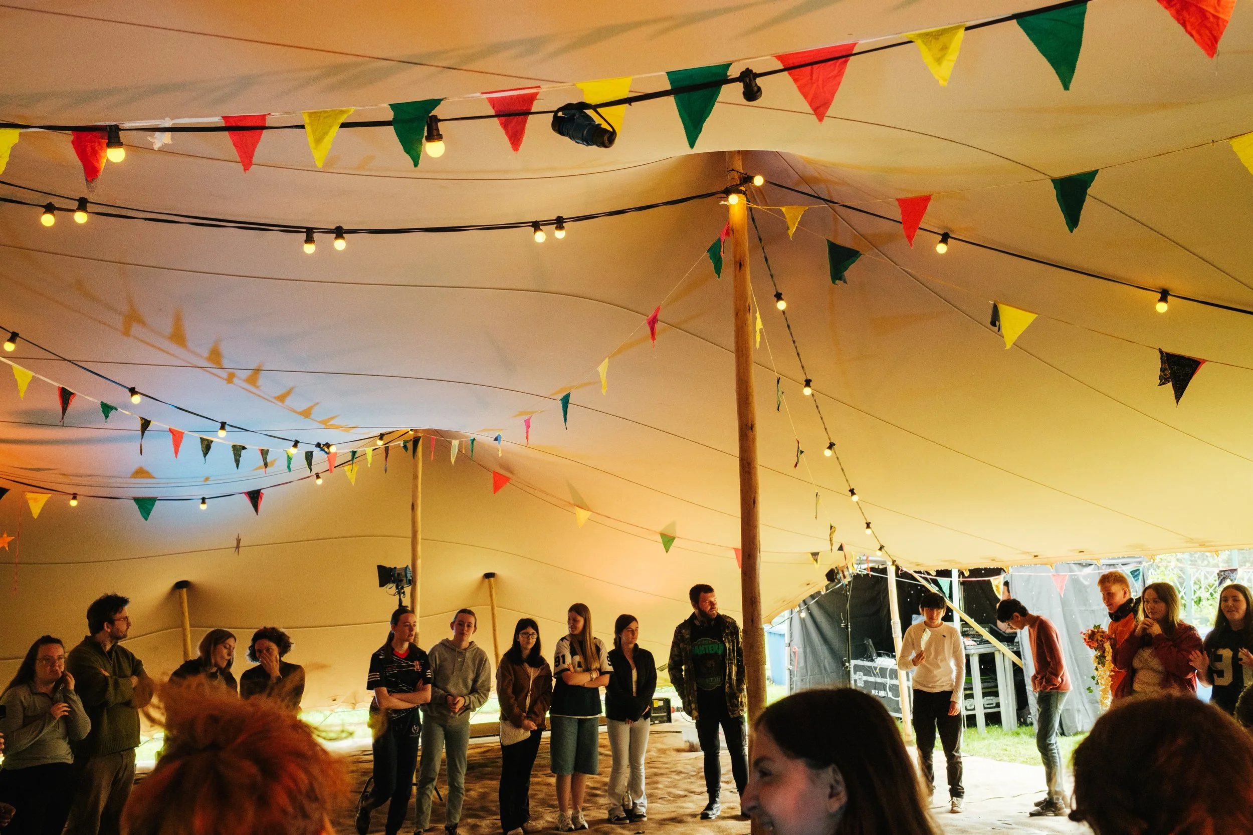 People standing inside a large tent decorated with colorful triangular pennant banners and string lights, possibly at a festival or celebration.