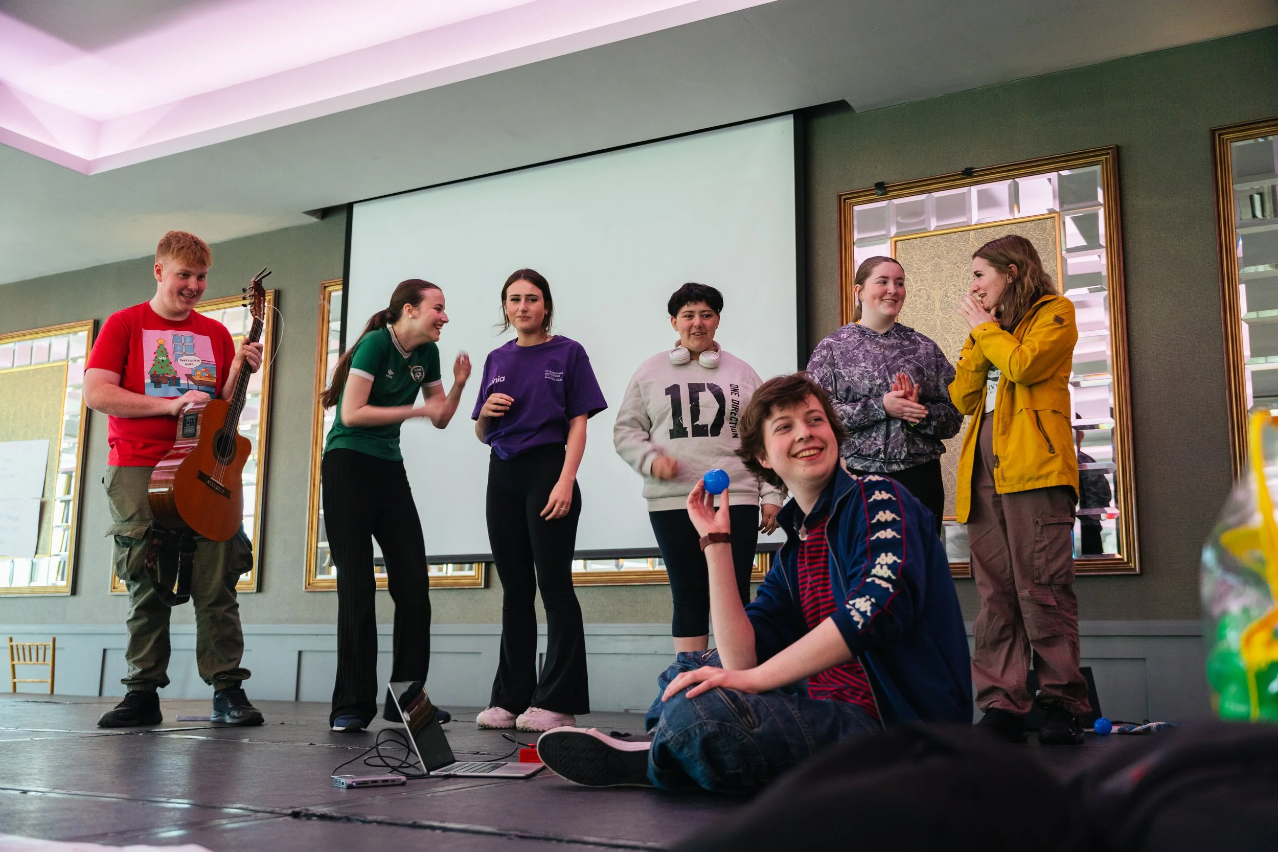 Group of seven young people on stage, some standing and one sitting, engaged in conversation and enjoying themselves, with a large screen behind them in a decorated room.