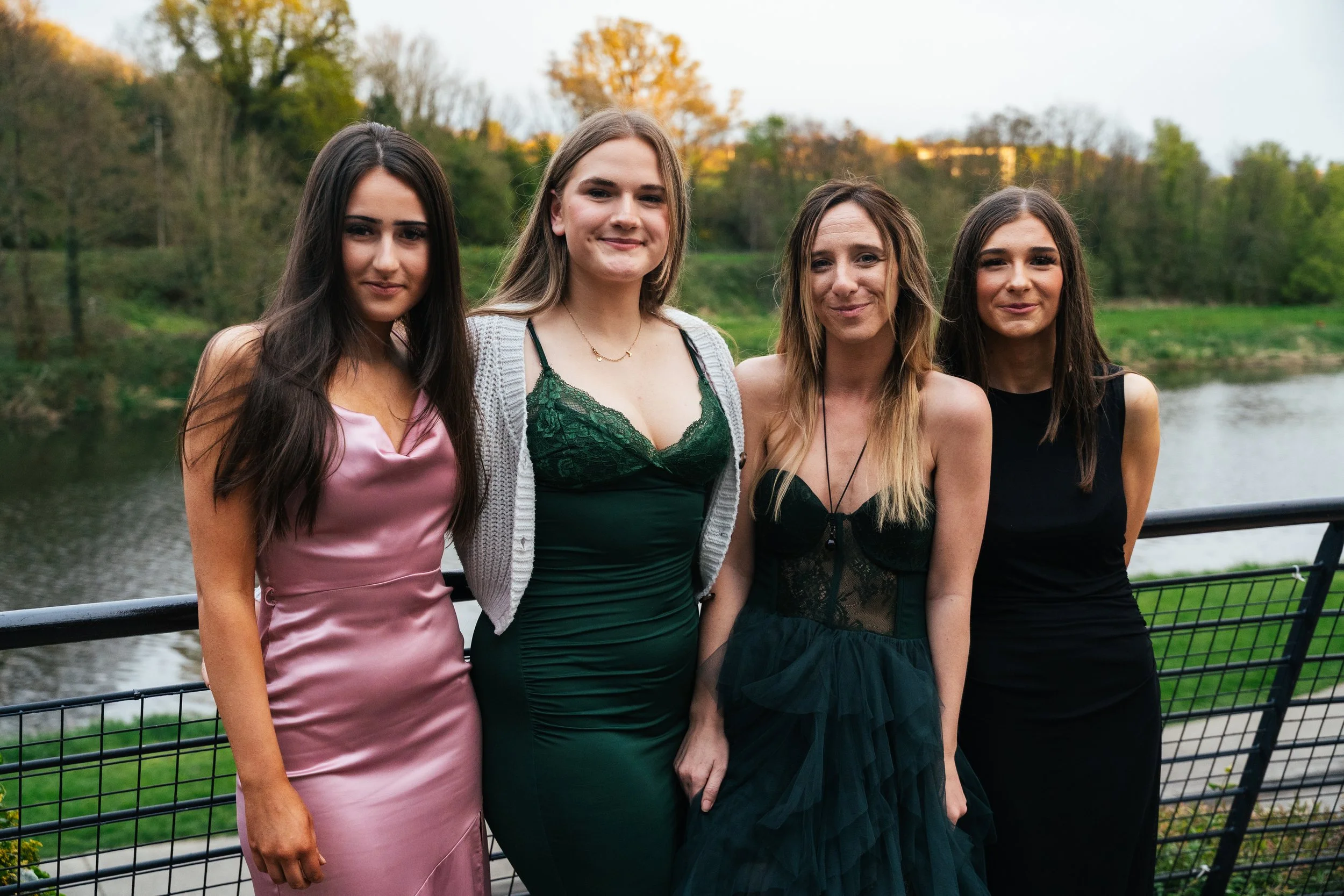 Four women in evening dresses standing outdoors near a body of water with trees in the background.
