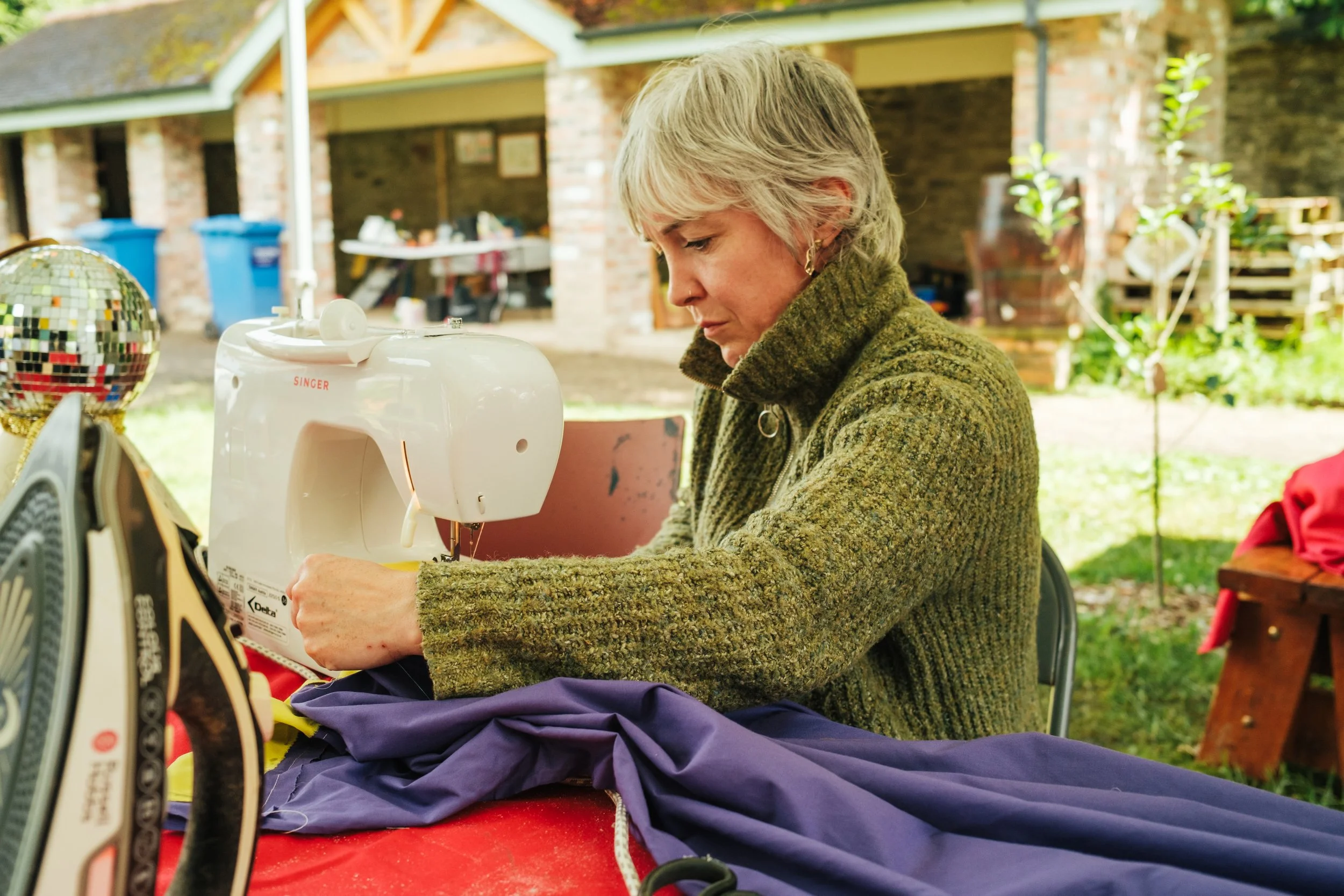 A woman sewing with a sewing machine outdoors, seated at a red table with fabric, in a backyard setting.